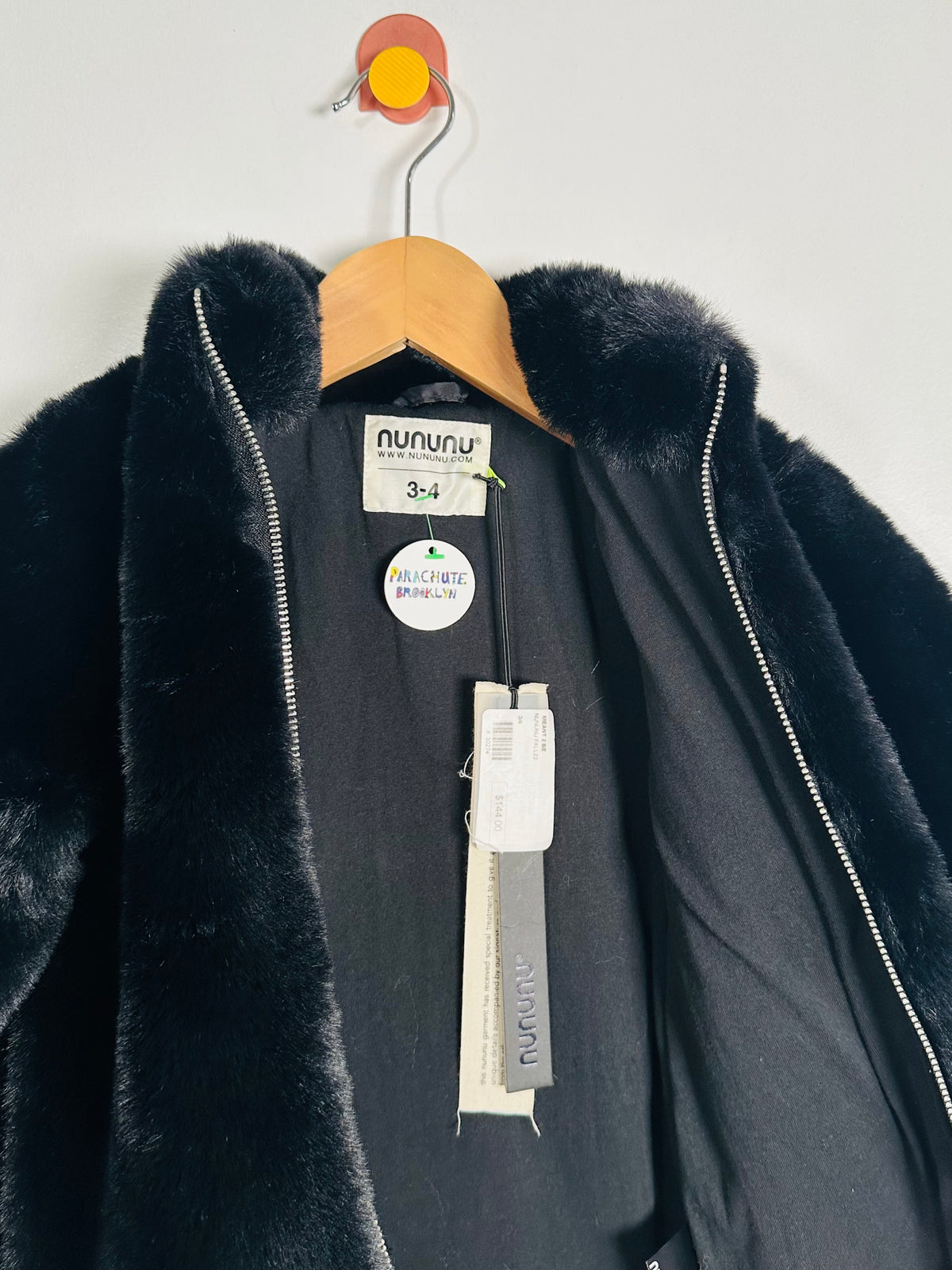 A black faux fur coat with a high collar and silver zipper, hanging on a wooden hanger against a plain light gray wall. The brand label 'nununu' is visible inside the collar, along with size and care tags.