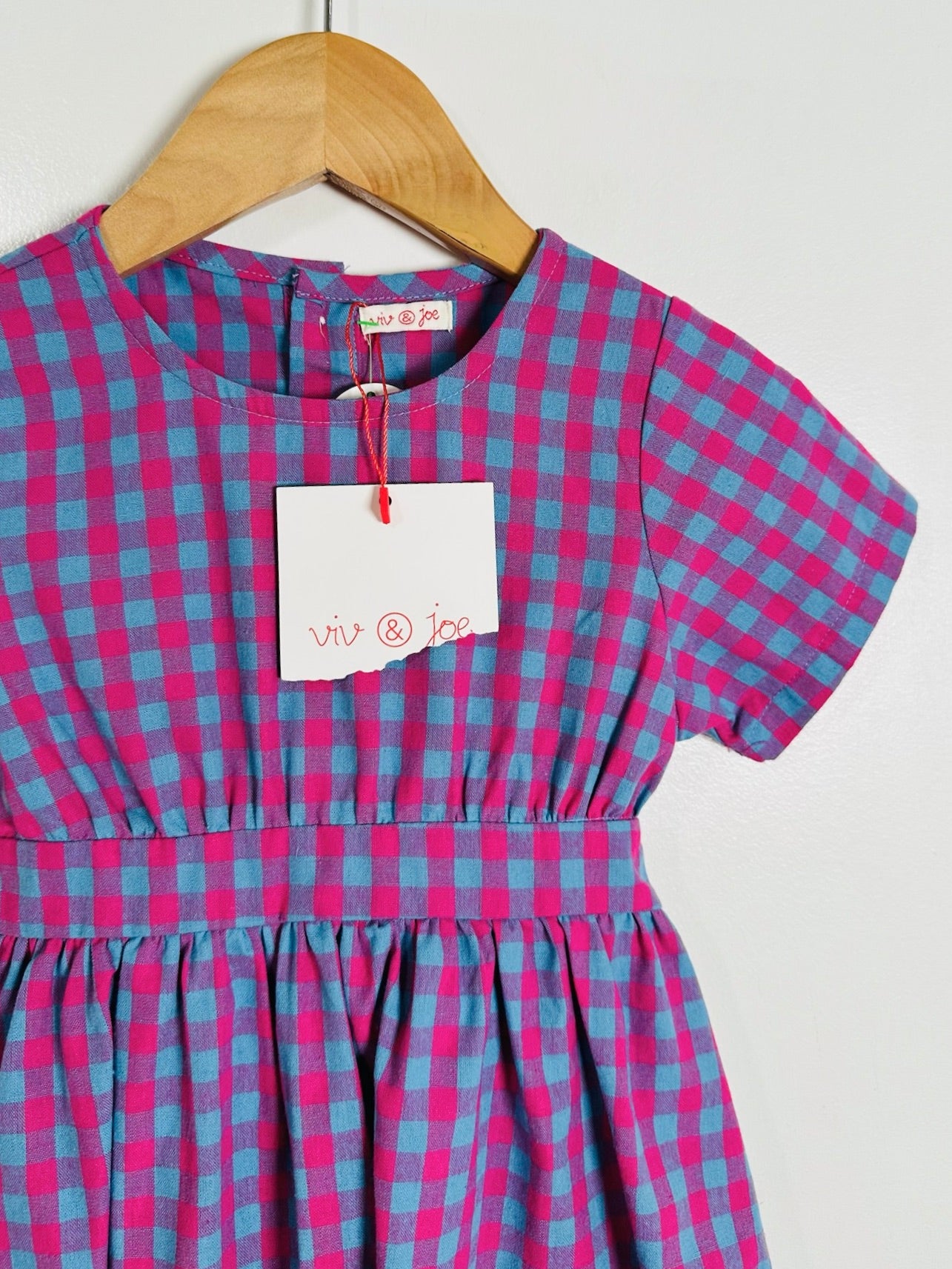 A short-sleeve children's dress with a pink and blue gingham check pattern, hanging on a wooden hanger against a plain light background. A white paper tag with red handwritten text is attached at the neckline.