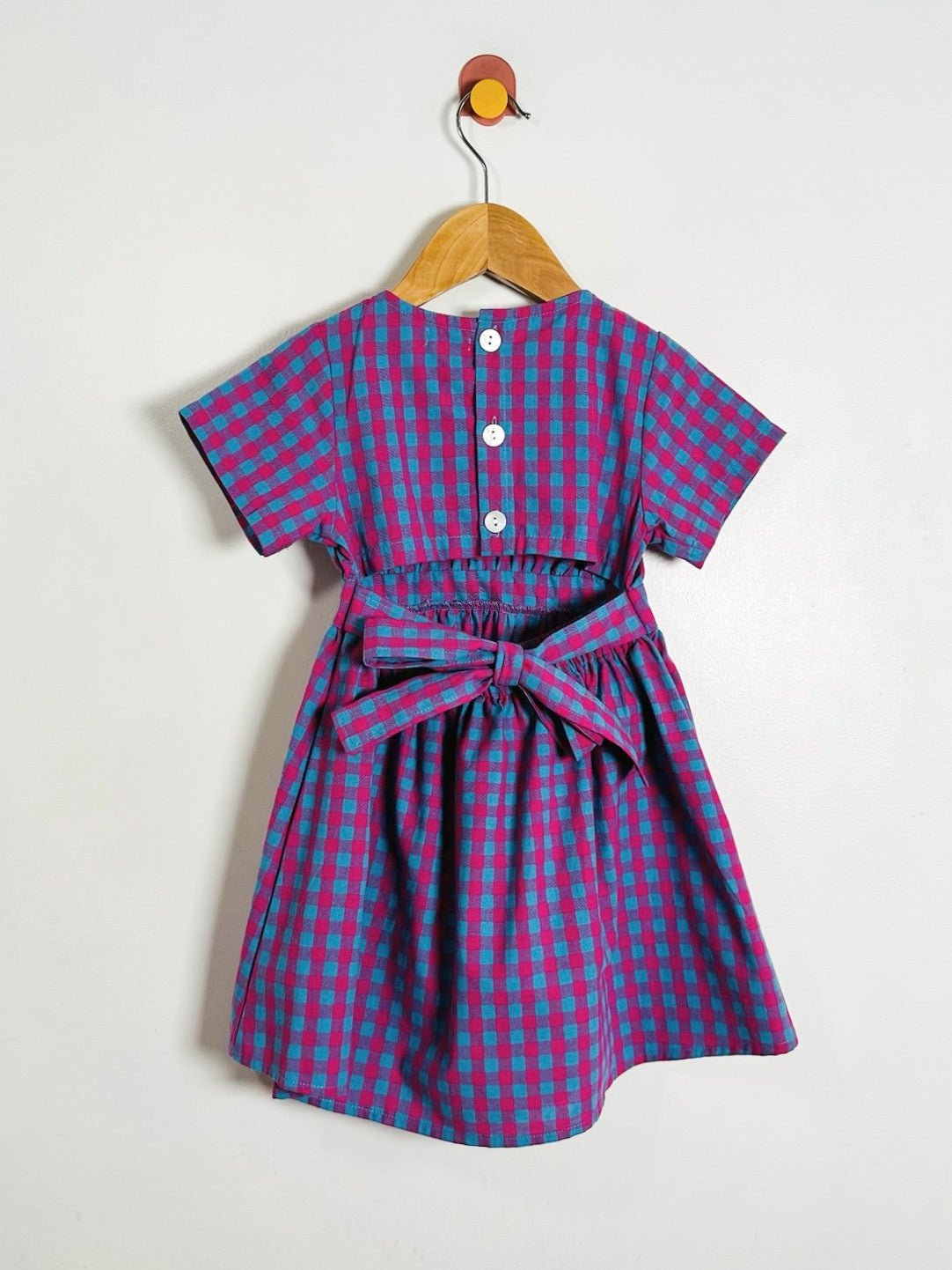 A short-sleeve children's dress with a pink and blue gingham check pattern, hanging on a wooden hanger against a plain white wall. The dress features a buttoned neckline, a gathered waist, and a matching bow tie at the waist.