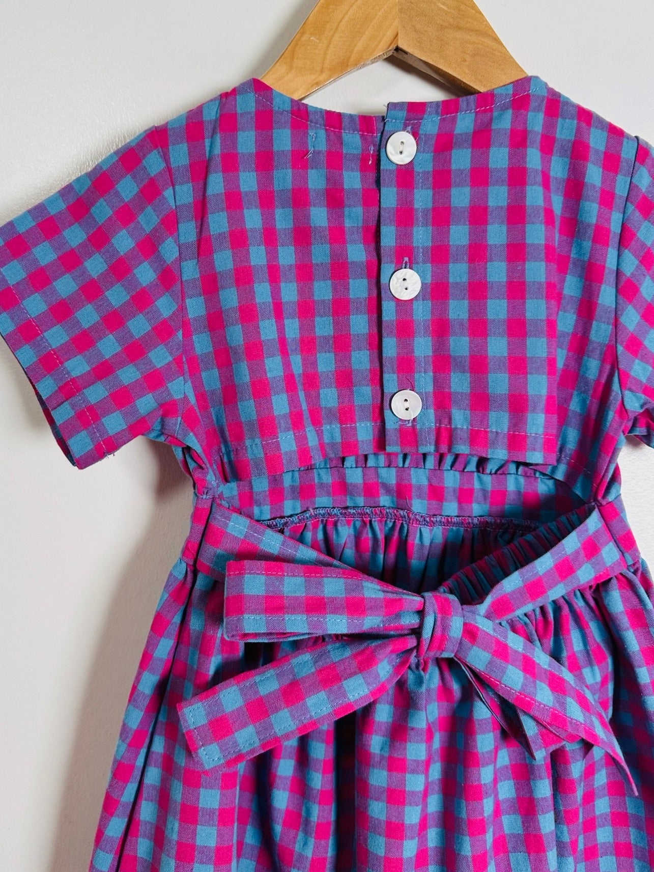 A short-sleeve children's dress with a pink and blue gingham check pattern, featuring a buttoned back and a matching fabric tie belt at the waist, hanging on a wooden hanger against a plain light background.