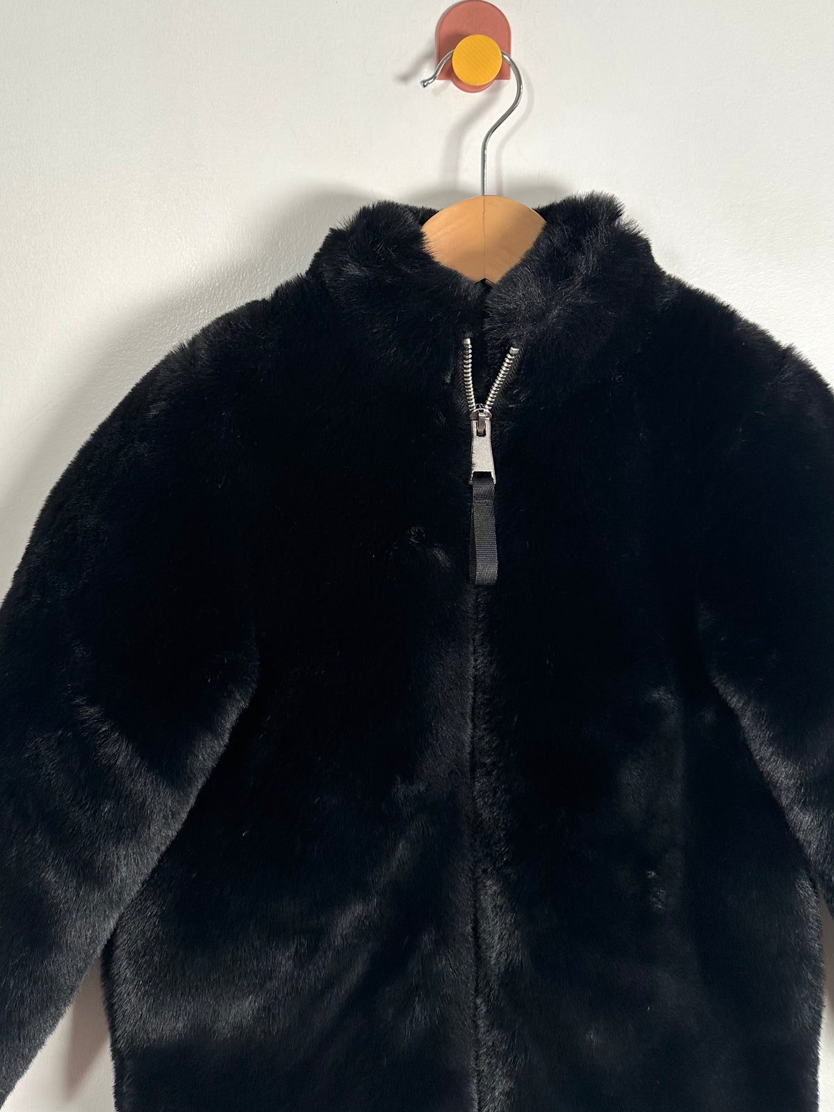 A black faux fur zip-up jacket with a high collar, hanging on a wooden hanger against a plain white wall.