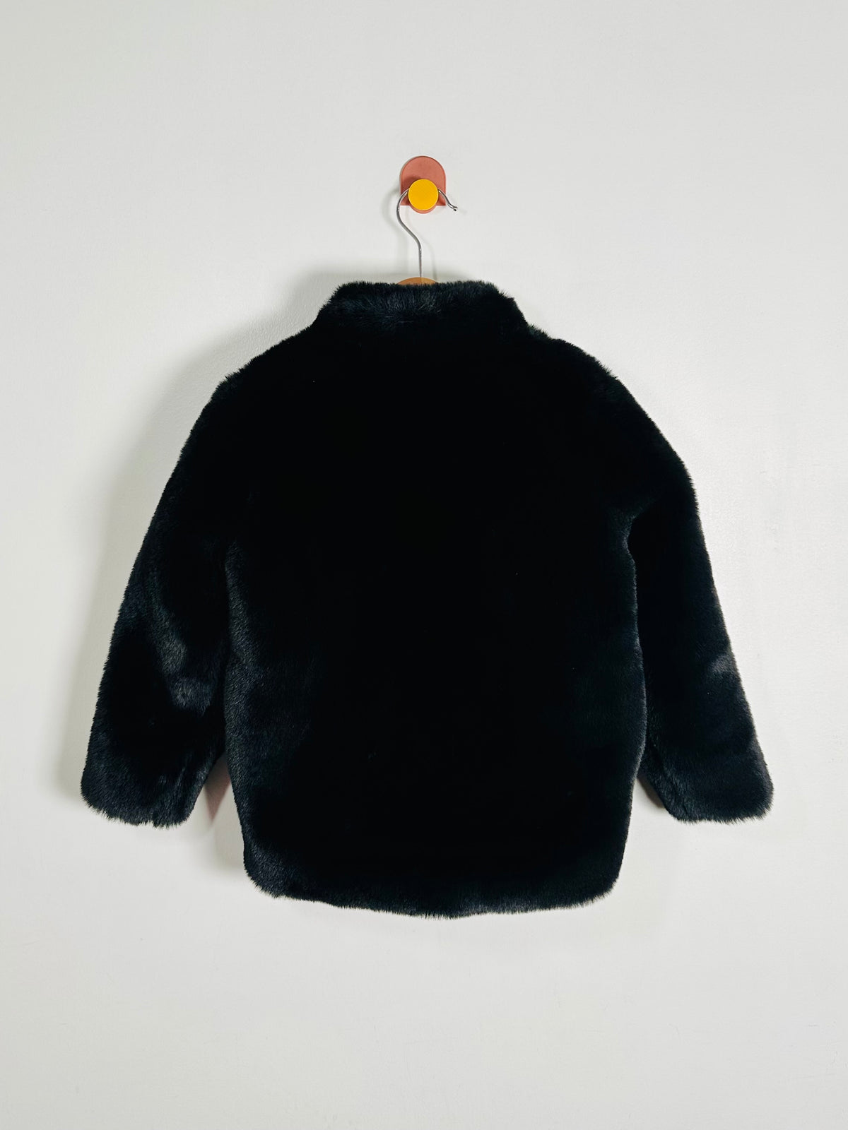 A black faux fur jacket with long sleeves and a high collar, shown from the back hanging on a wooden hanger against a plain white wall.
