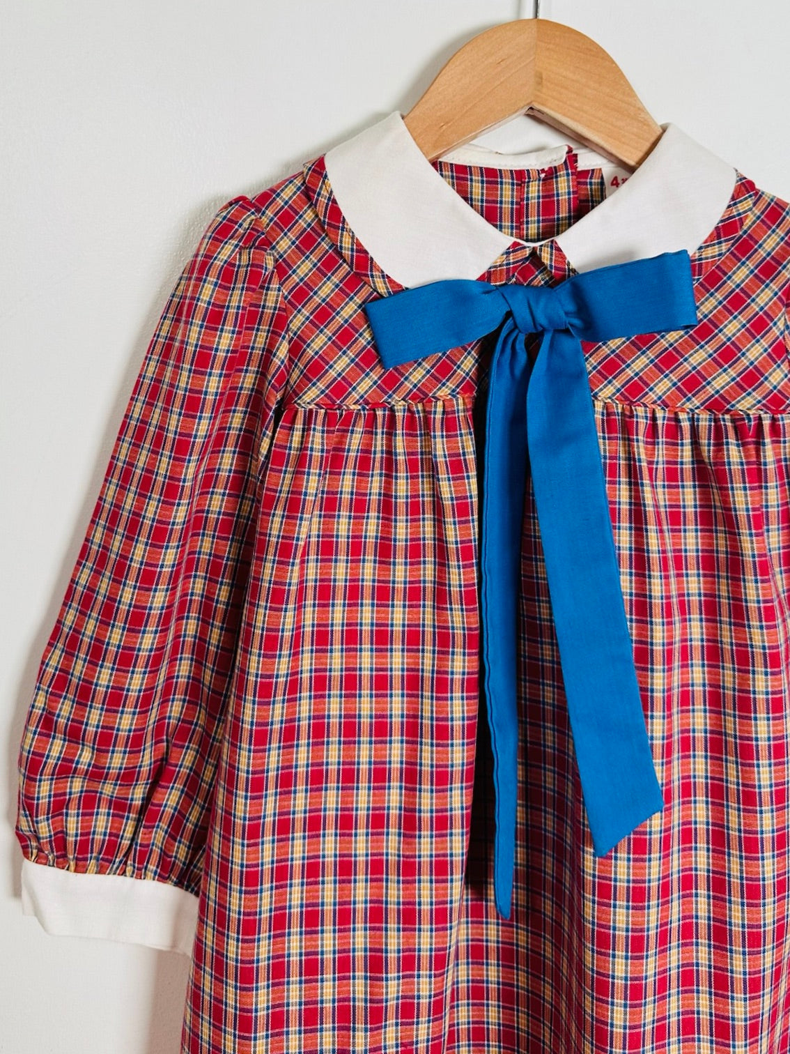 A children's long-sleeve plaid dress hanging on a wooden hanger against a plain light background. The dress has a red, yellow, and blue plaid pattern, a white collar, and a large blue bow at the neckline.