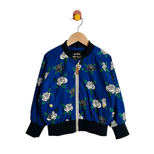 A blue bomber jacket with a white and black floral print, hanging on a wooden hanger against a plain white background.