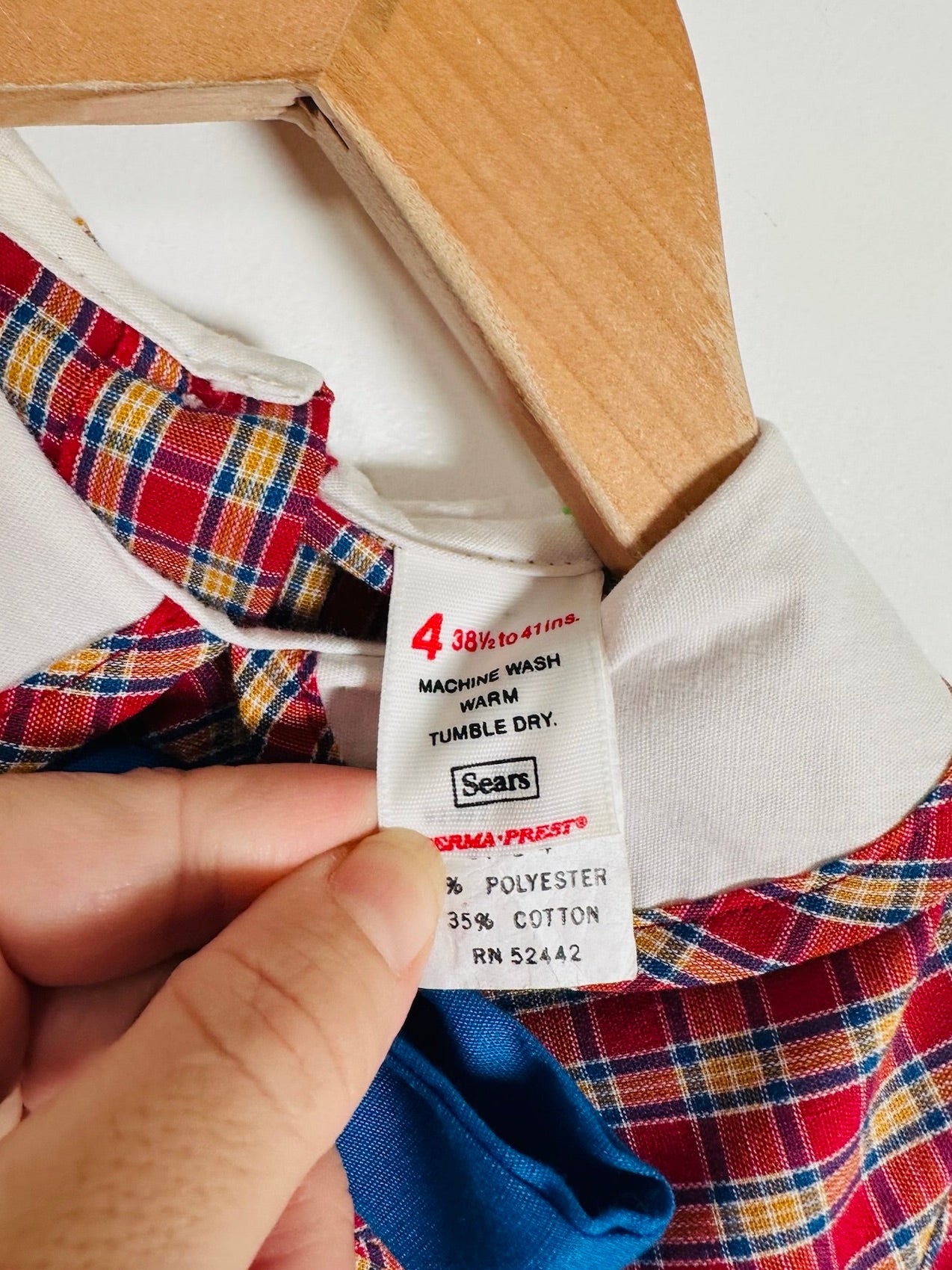Close-up of a clothing care label reading Sears and Serma-Frust's, held by a hand against a red, yellow, and blue plaid garment on a wooden hanger.