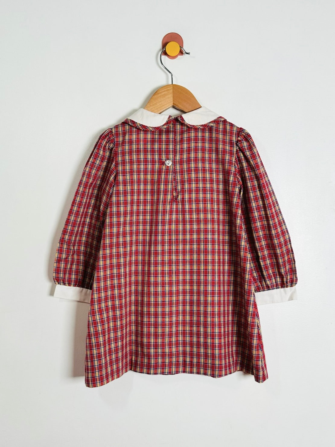 A red and green plaid long-sleeve shirt dress with a white Peter Pan collar, hanging on a wooden hanger against a plain white wall.