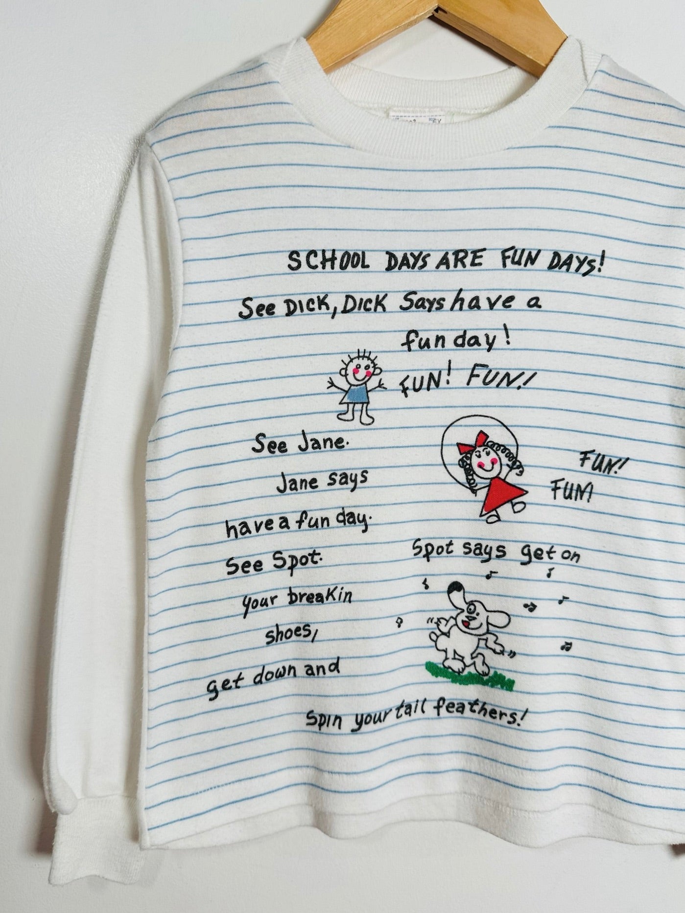 A white long-sleeve t-shirt with light blue horizontal lines, hanging on a wooden hanger against a white wall. The shirt features a black hand-drawn style graphic with text reading 'SCHOOL DAYS ARE FUN DAYS!' and cartoon doodles.