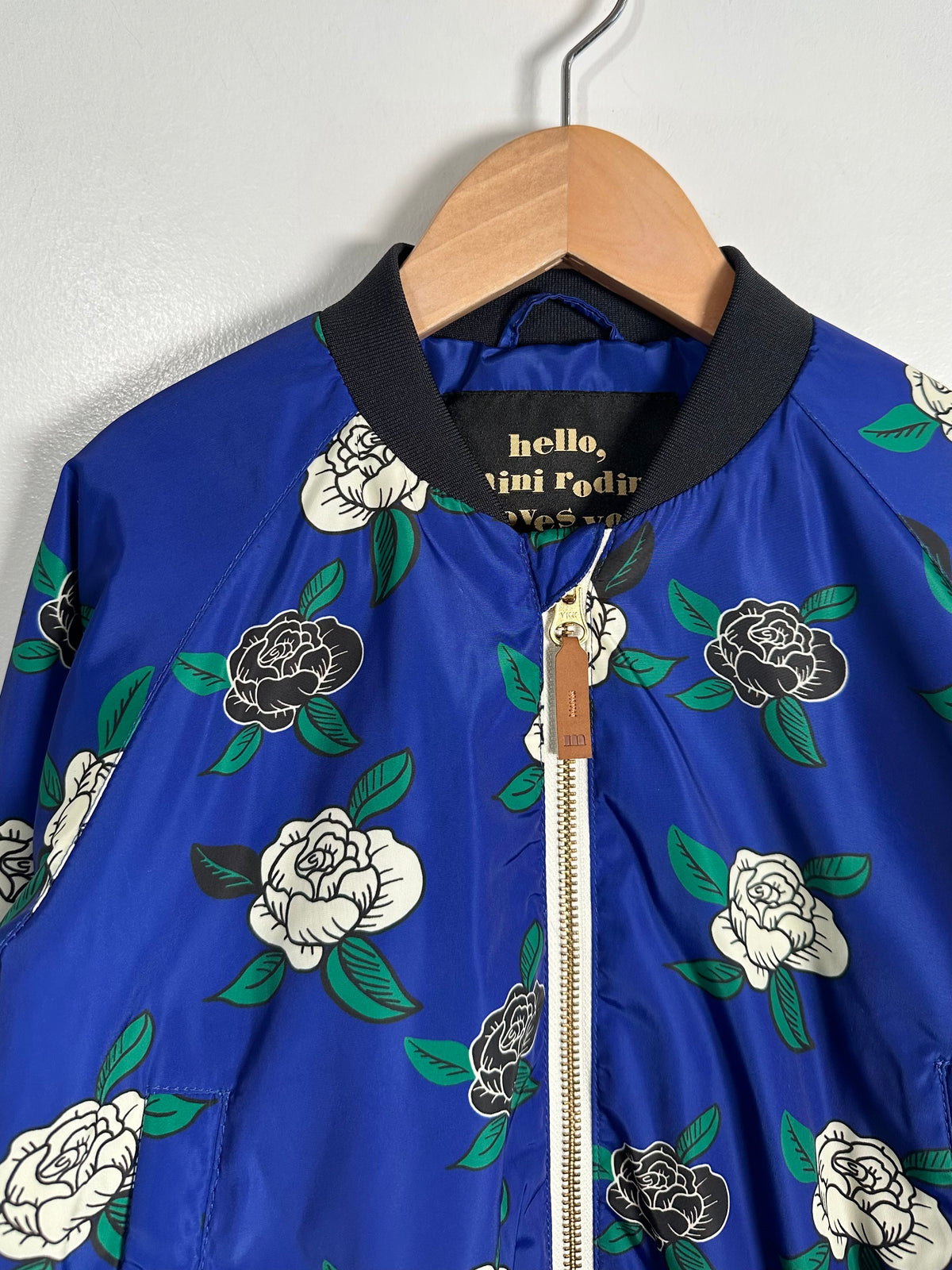 A close-up of a vibrant royal blue bomber jacket featuring a black, white, and green floral print, hanging on a wooden hanger against a light gray wall. The brand name 'hello, vini Fodir' is visible on the inner neck label.