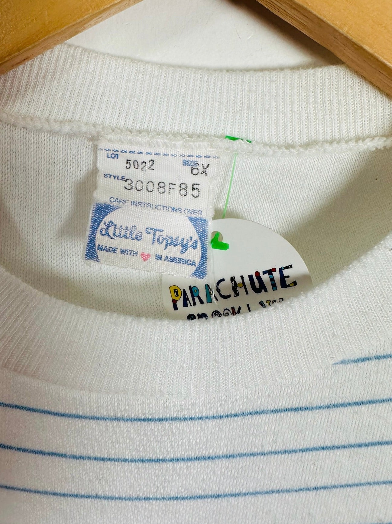 Close-up of a white ribbed knit garment with thin blue horizontal stripes, showing the inner neck labels including Little Topy's and a PARACHUTE brand tag.