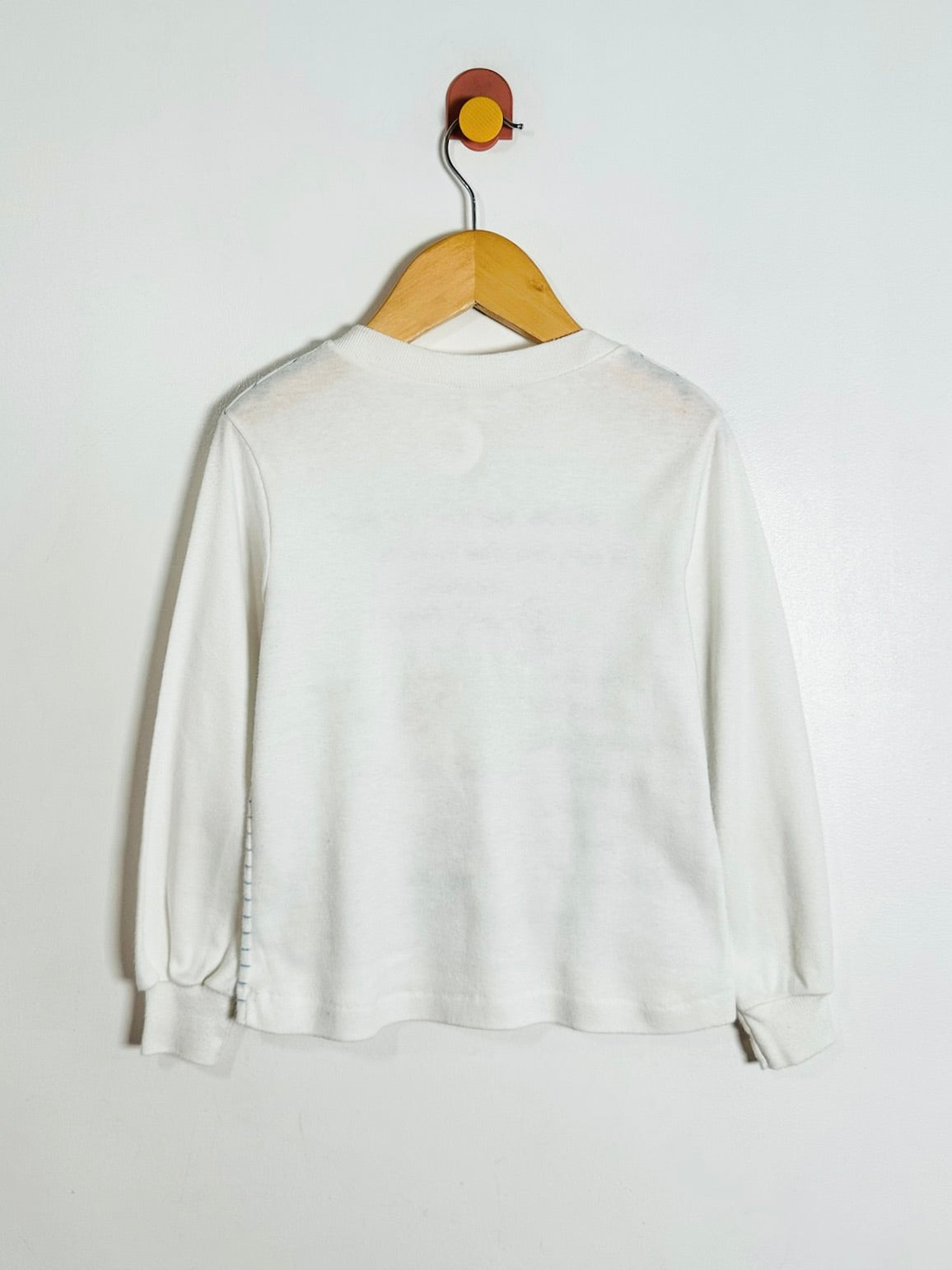 A white long-sleeve crewneck top hanging on a wooden hanger against a plain white wall.