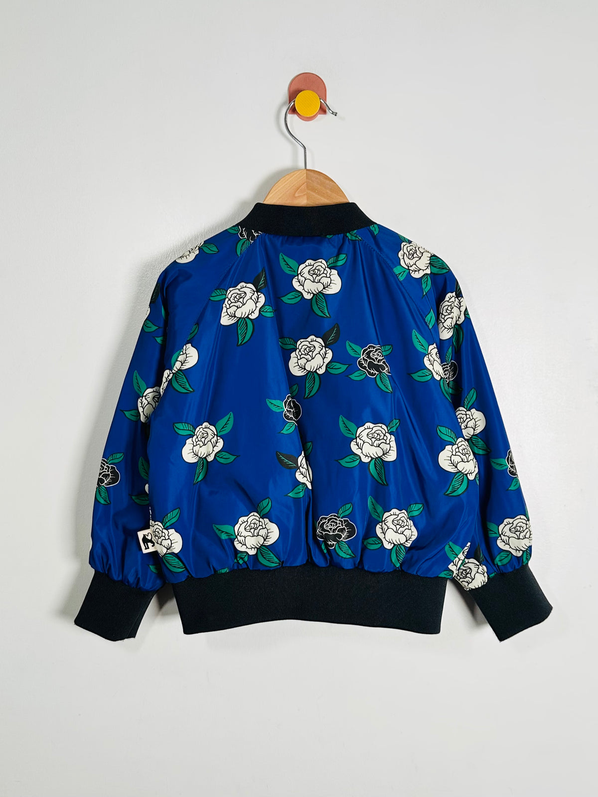 A blue satin bomber jacket with a white rose and green leaf pattern, hanging on a wooden hanger against a plain light gray wall.