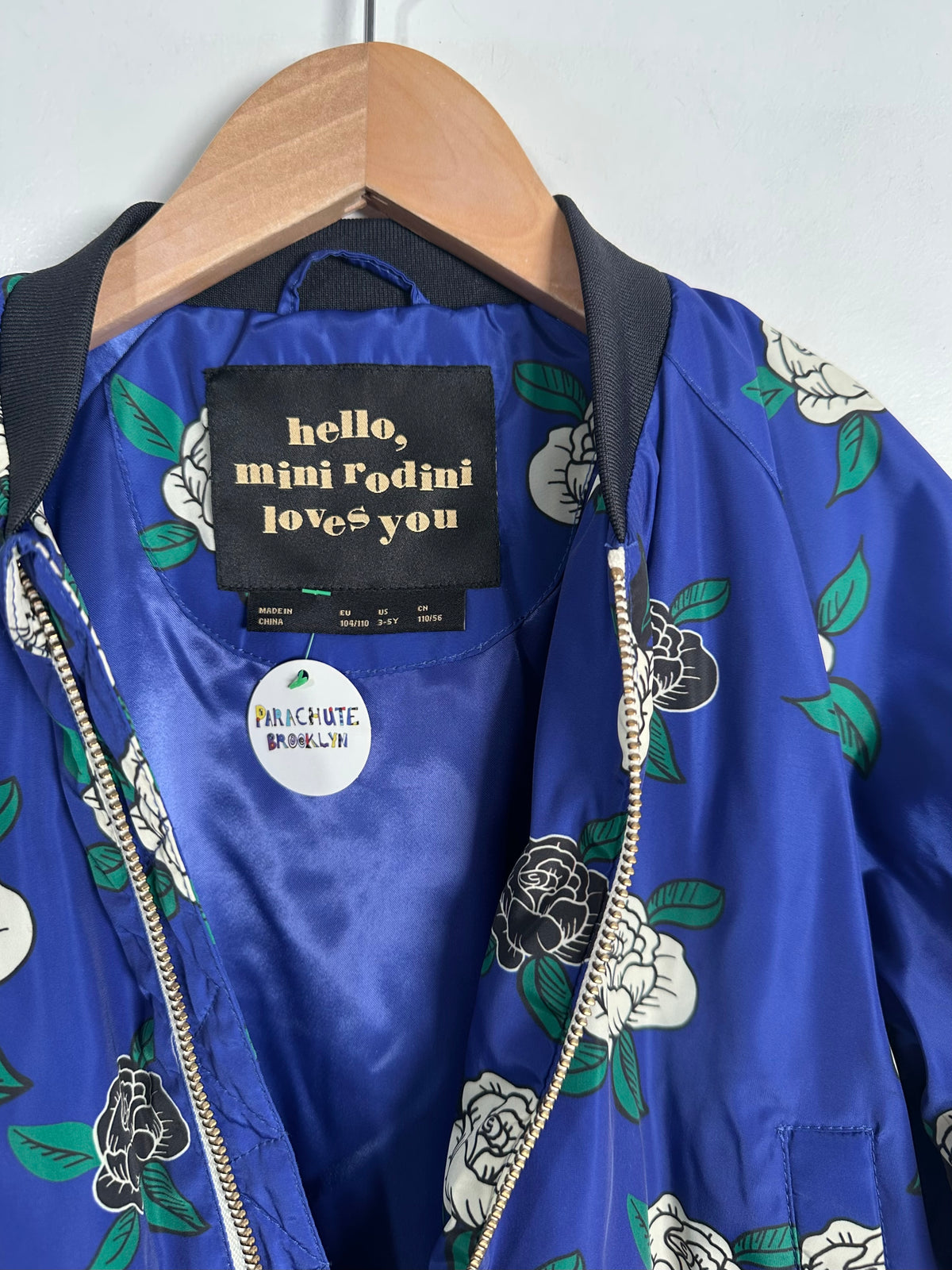 A close-up of a blue satin bomber jacket hanging on a wooden hanger against a light gray wall. The jacket features a white and green floral print, a black collar, and a black interior label that reads 'hello, mini Rodini love you' with a visible 'PUNCHUTE BLOOM' sticker on the inside.