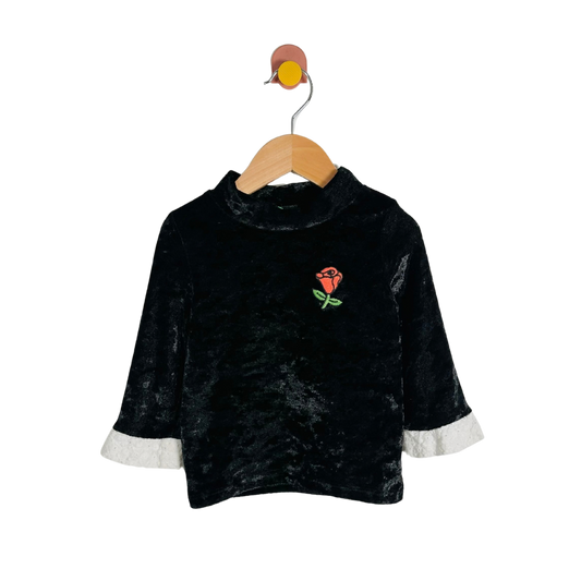 A black velvet long-sleeve top with a mock neck, featuring a small embroidered red rose on the chest and contrasting white cuffs, hanging on a wooden hanger against a white background.