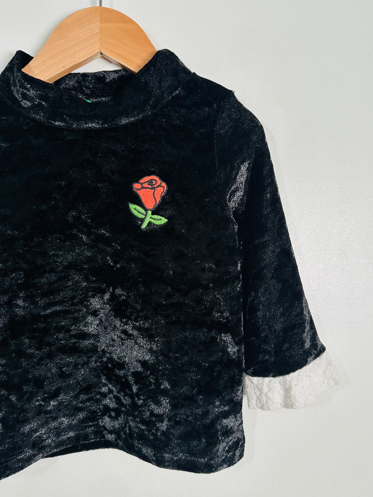 A black velvet long-sleeve top with a white lace cuff and an embroidered red rose on the chest, hanging on a wooden hanger against a plain light gray wall.