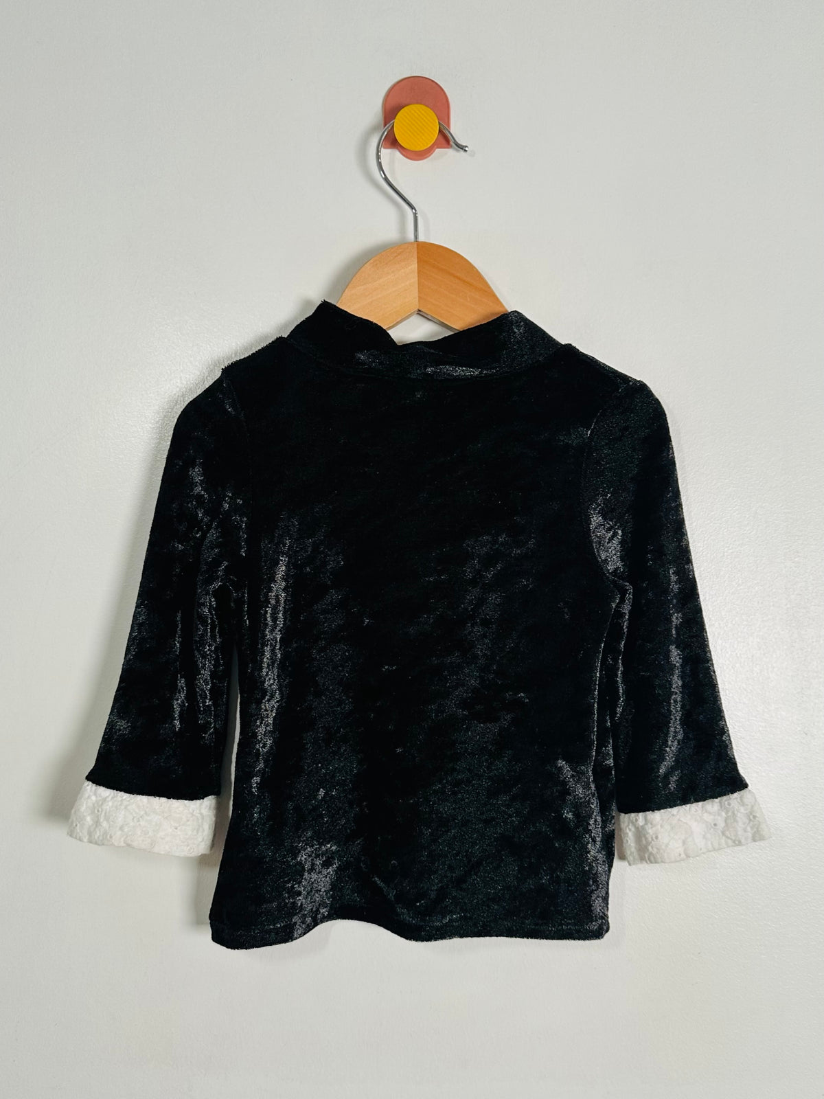 A black velvet long-sleeve top with white faux-fur trimmed cuffs, hanging on a wooden hanger against a plain light gray wall.