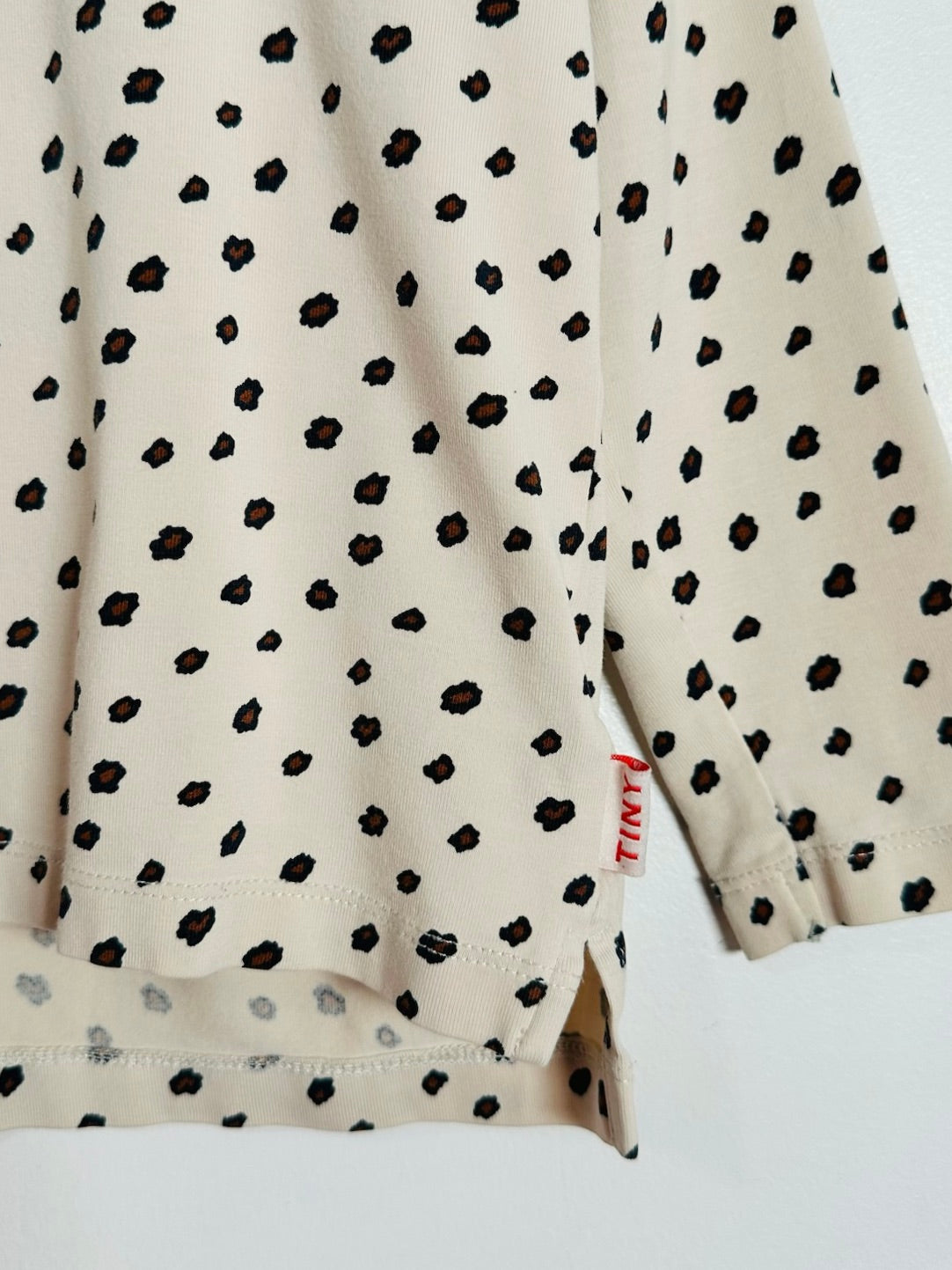 Close-up of a long-sleeve cream-colored top with a black spotted pattern, featuring a small red 'LUMI' brand tag near the cuff.
