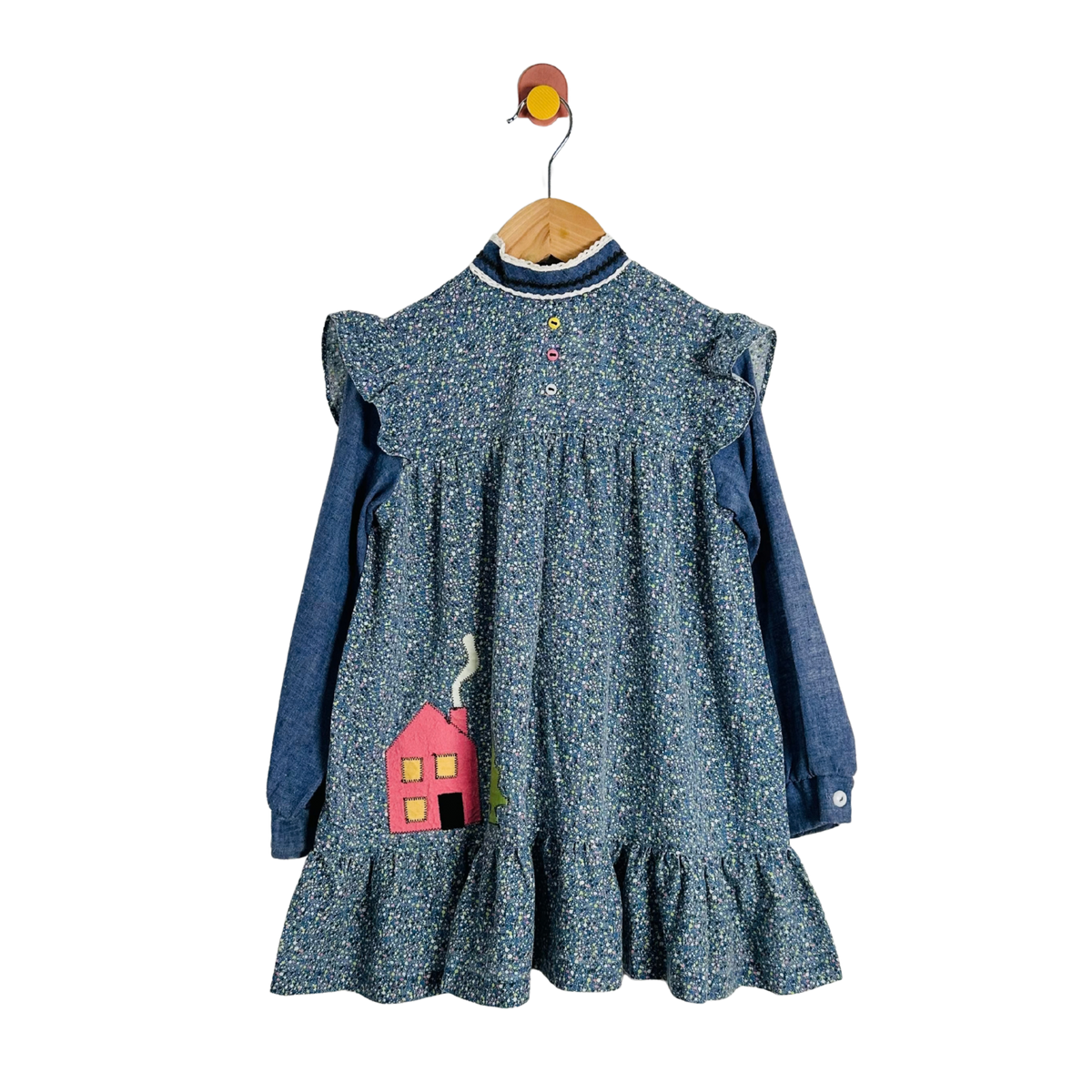 A children's long-sleeve dress with a blue and white floral print over a solid blue base, hanging on a wooden hanger with a bird hook. The dress features ruffled shoulder details, a buttoned neckline, and a pink house applique on the front.