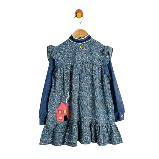 A children's long-sleeve dress with a blue and white floral print over a solid blue base, hanging on a wooden hanger with a bird hook. The dress features ruffled shoulder details, a buttoned neckline, and a pink house applique on the front.