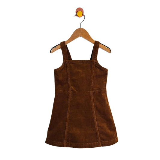 A brown corduroy pinafore dress with wide shoulder straps, hanging on a wooden hanger against a plain white background.