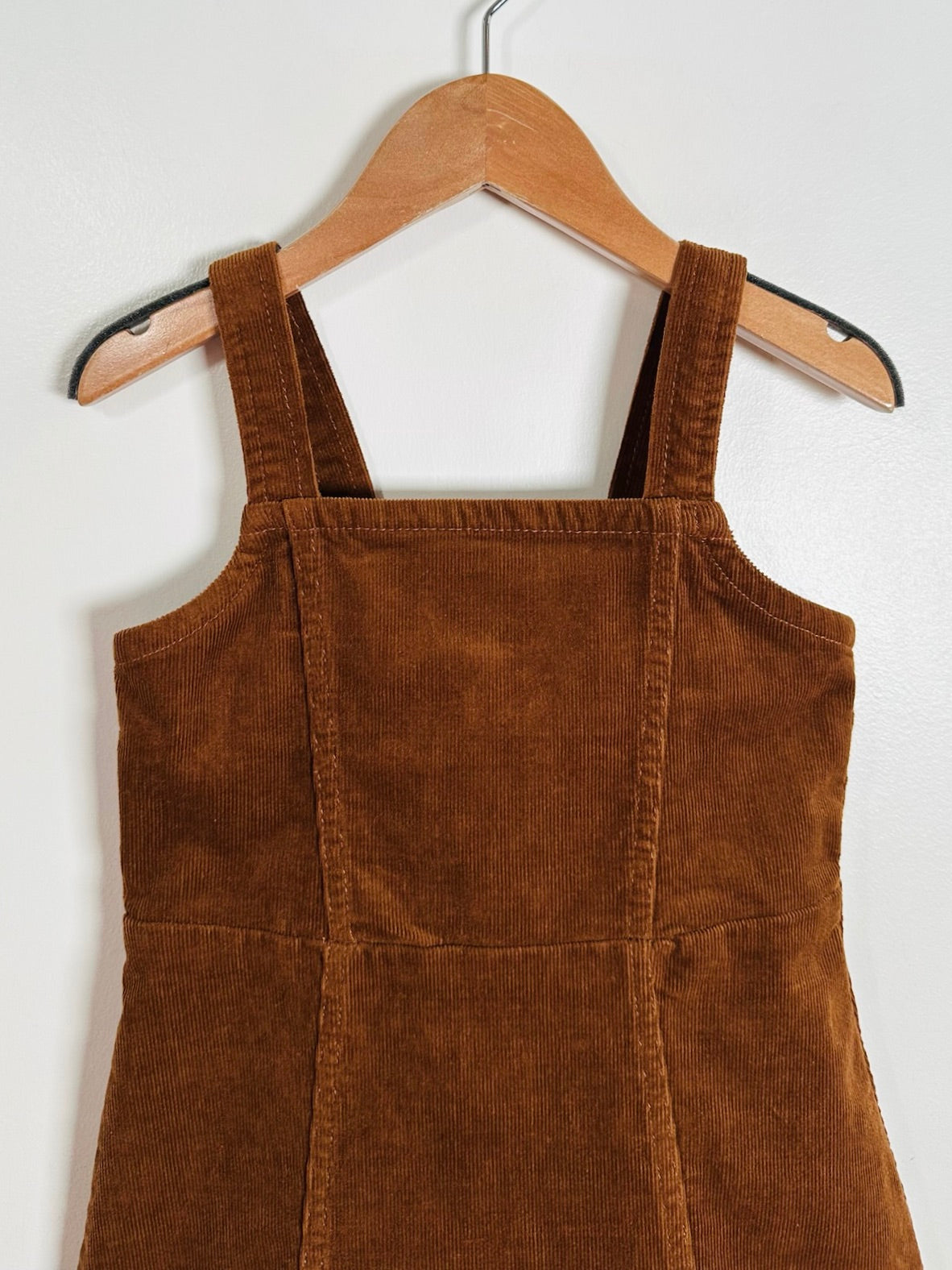 A brown corduroy pinafore dress with wide shoulder straps, hanging on a wooden hanger against a plain white wall.