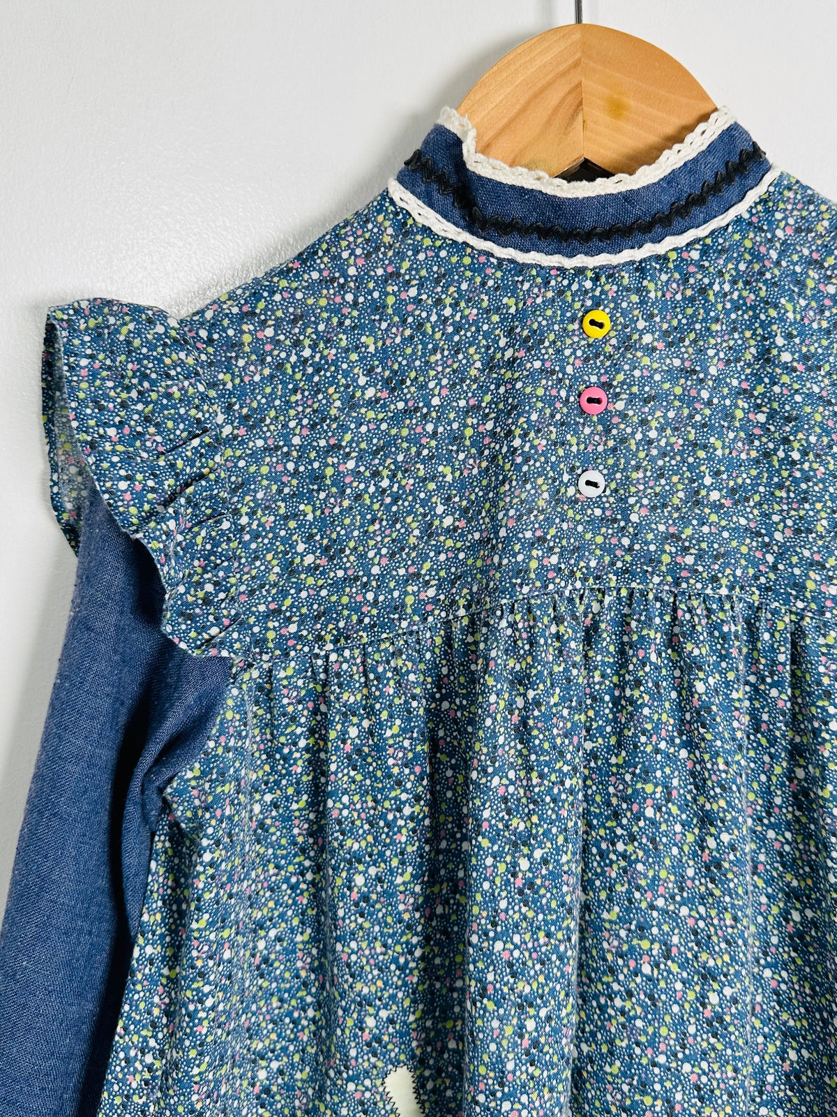 A blue floral long-sleeve dress with a smocked bodice and ruffled collar, hanging on a wooden hanger against a light gray wall.