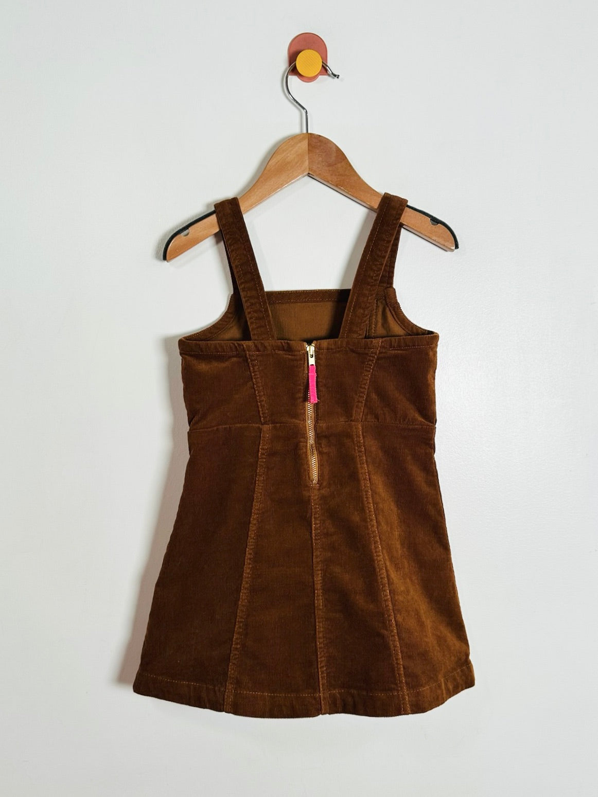 A brown corduroy pinafore dress with a front zipper, hanging on a wooden hanger against a plain white wall.