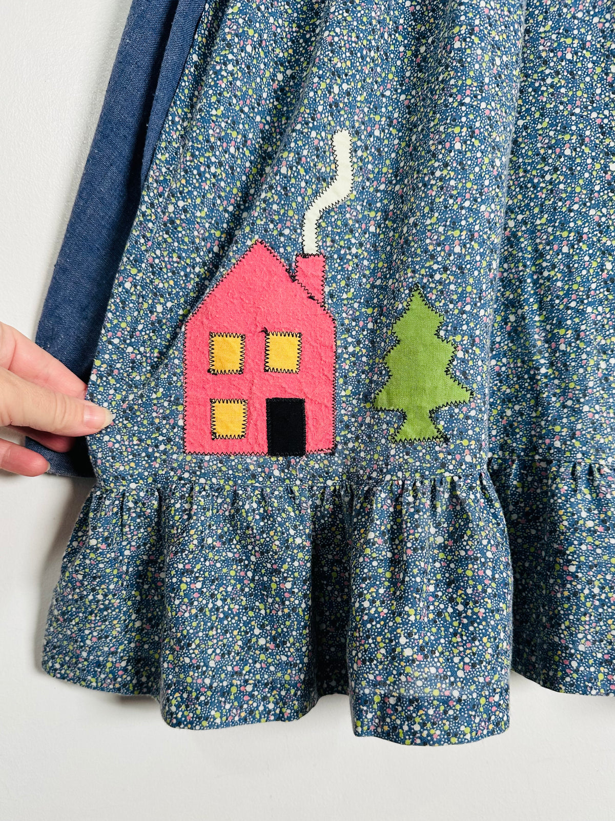 Close-up of a blue floral skirt with a pink house and green tree applique on the front, held by a hand on the left side.