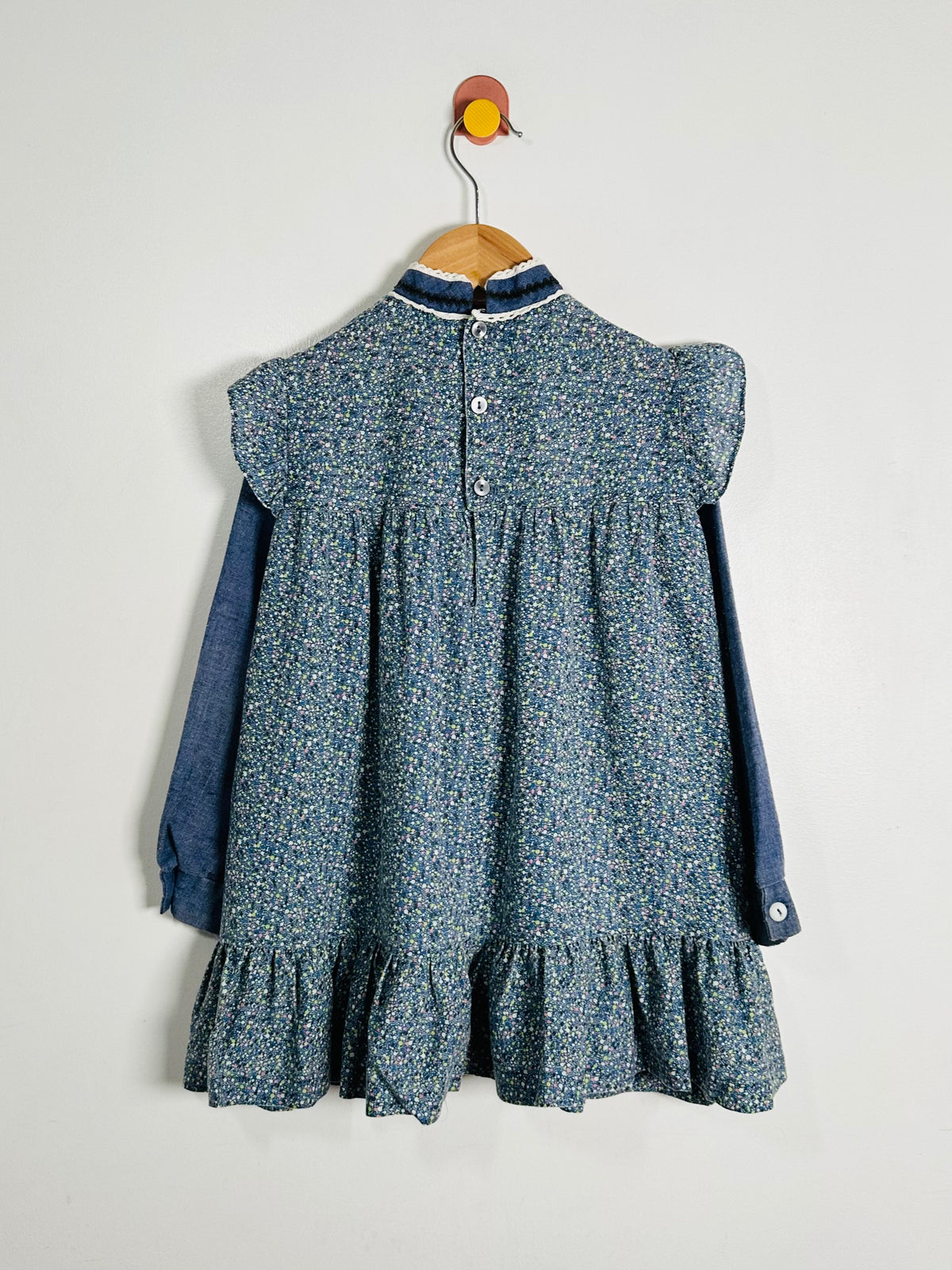 A sleeveless baby dress with a blue and white floral print, featuring a ruffled hem, short puff sleeves, and a buttoned back, layered over a solid blue long-sleeve shirt. The dress is hanging on a wooden hanger from a wall hook against a plain light gray wall.