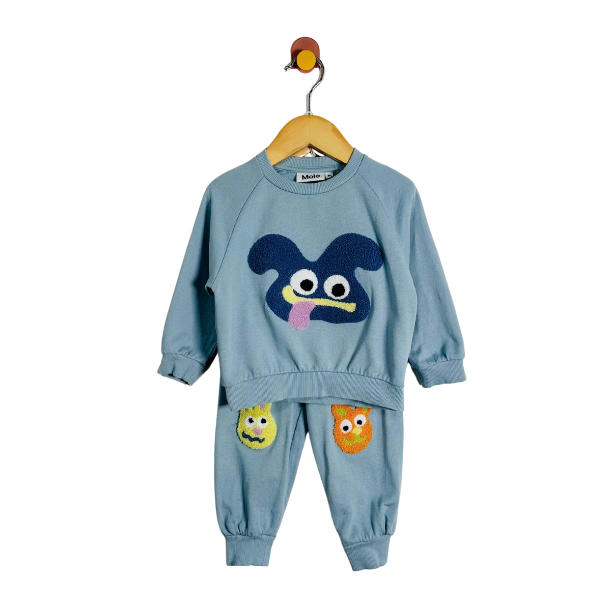 A toddler two-piece sweatshirt and jogger set in dusty blue, hanging on a wooden hanger. The set features a large dark blue monster face applique on the front of the sweatshirt, matching blue pants with smaller yellow and orange monster face appliques on the thighs, and a small bird-shaped hanger hook at the top.