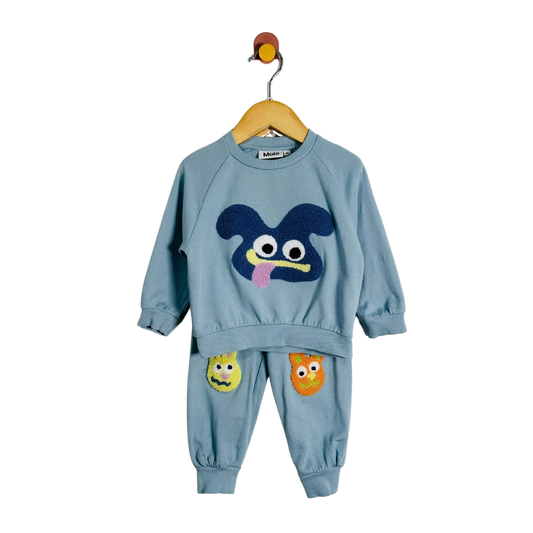 A toddler two-piece sweatshirt and jogger set in dusty blue, hanging on a wooden hanger. The set features a large dark blue monster face applique on the front of the sweatshirt, matching blue pants with smaller yellow and orange monster face appliques on the thighs, and a small bird-shaped hanger hook at the top.