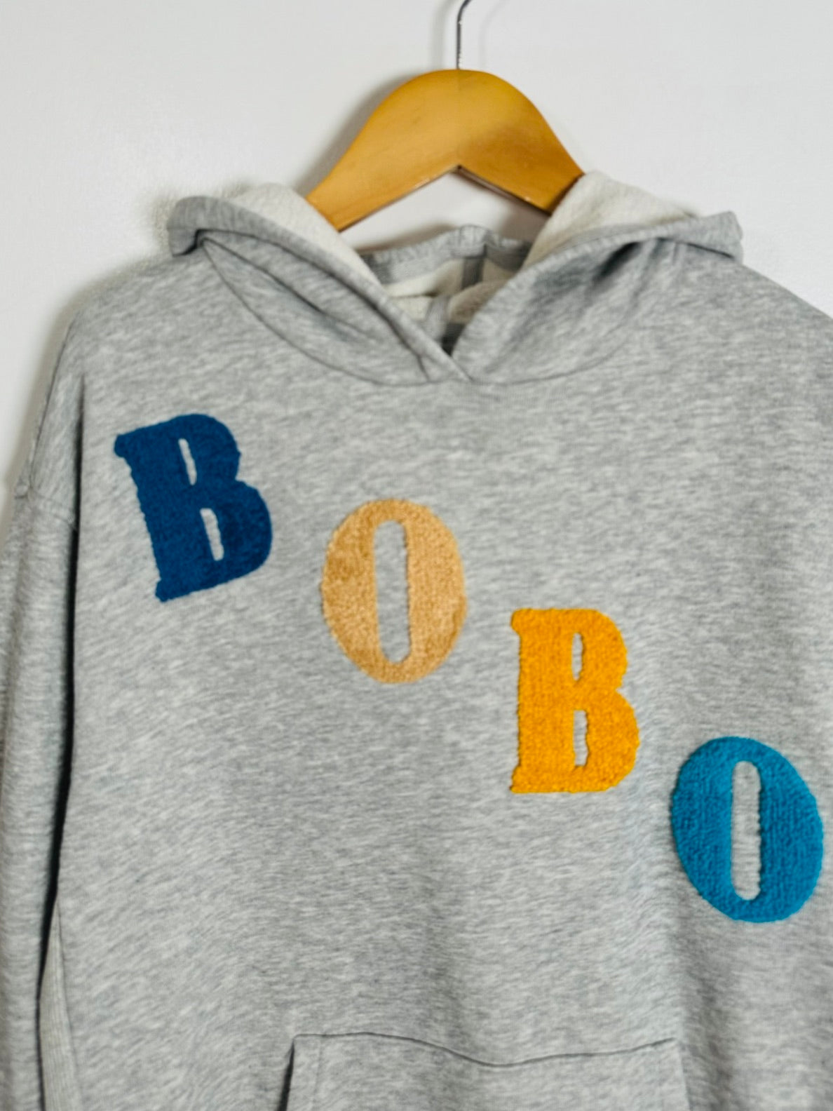 A heather gray pullover hoodie hanging on a wooden hanger against a plain white wall. The front features the word 'BOBO' spelled out in large, textured letters in blue, mustard yellow, and teal.
