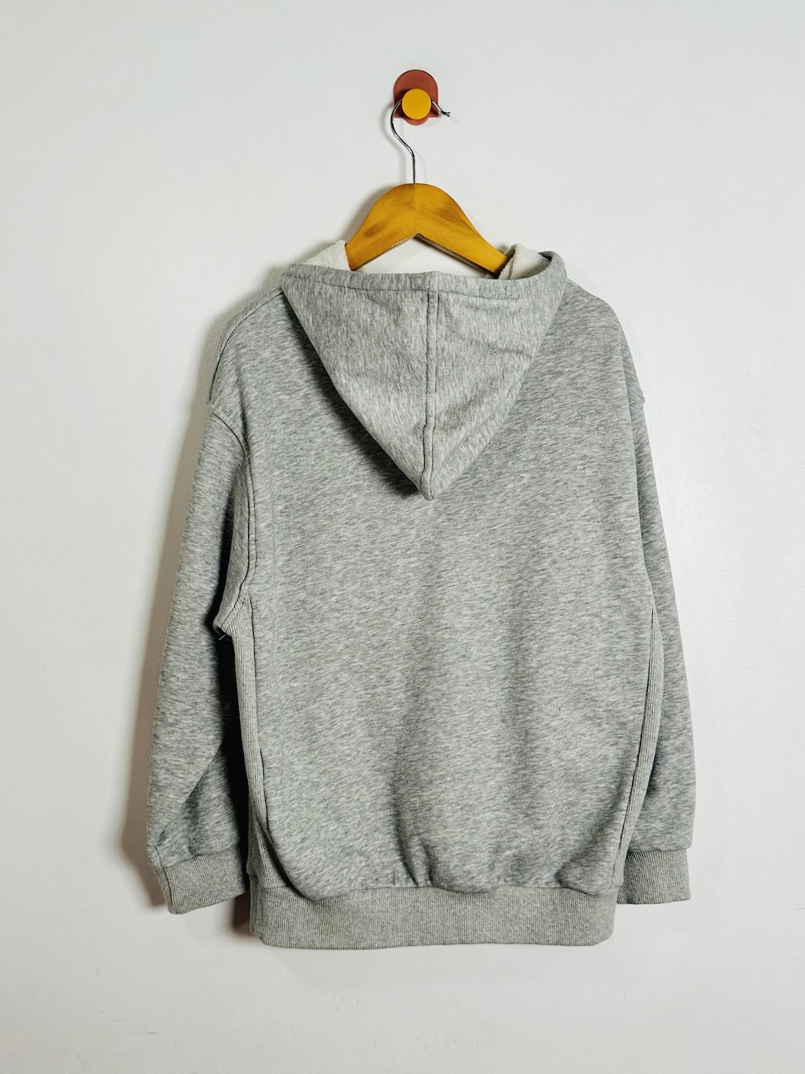 A heather gray pullover hoodie shown from the back, hanging on a wooden hanger against a plain off-white wall.