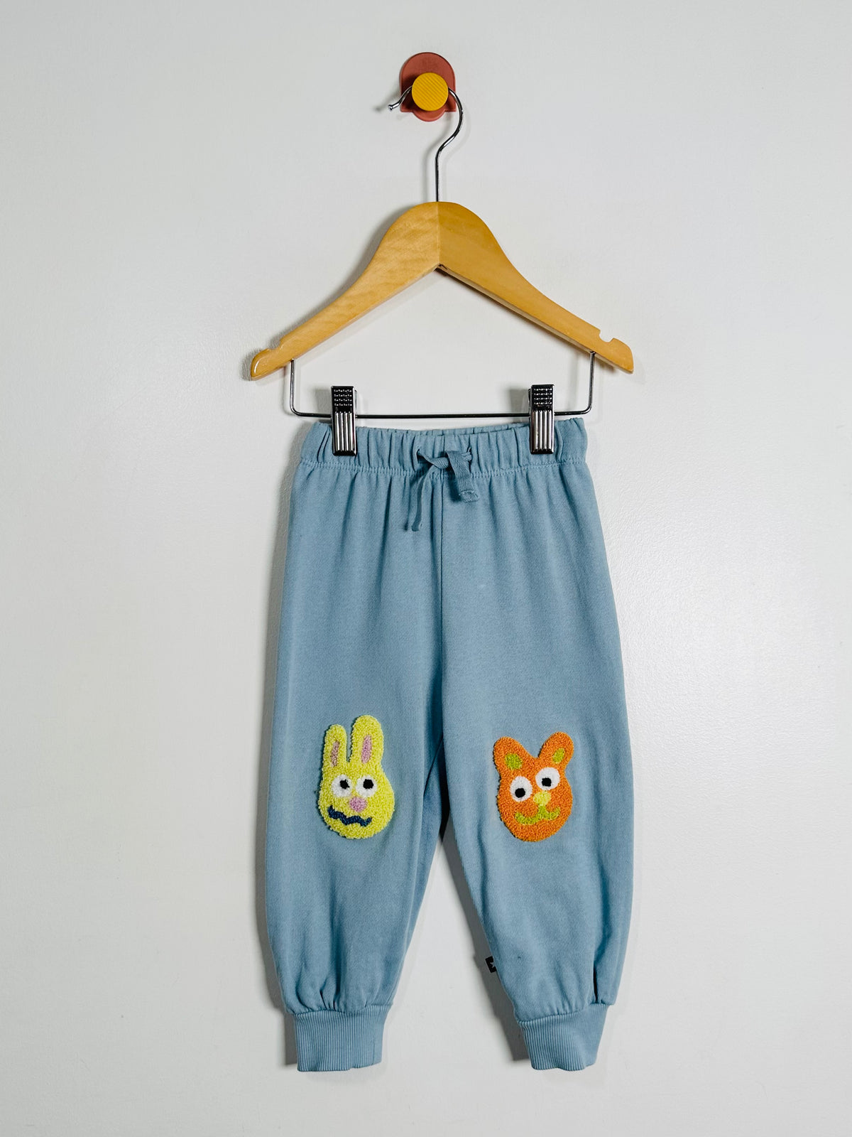 Light blue jogger pants with embroidered bunny and owl faces, hanging from a wooden hanger against a plain light gray wall.
