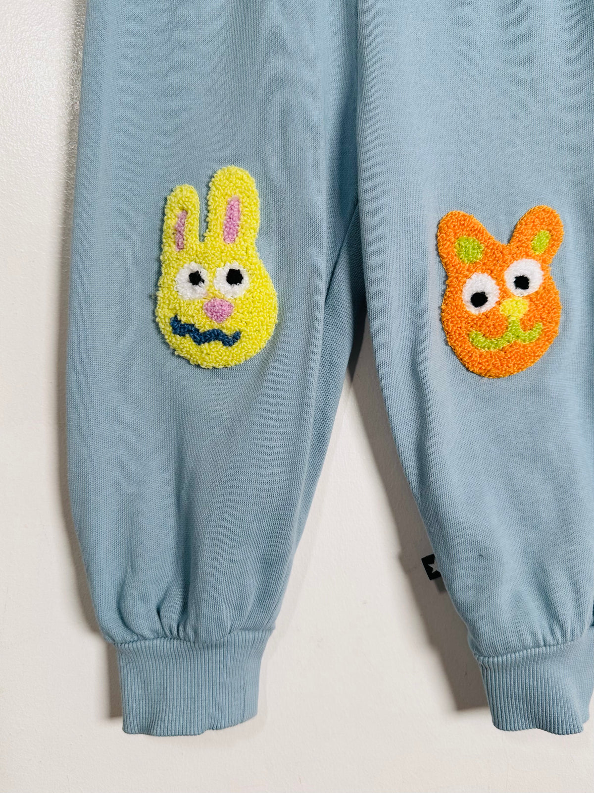 Light blue sweatpants with two colorful, raised fuzzy bunny and cat face patches on the thighs, shown against a plain off-white background.