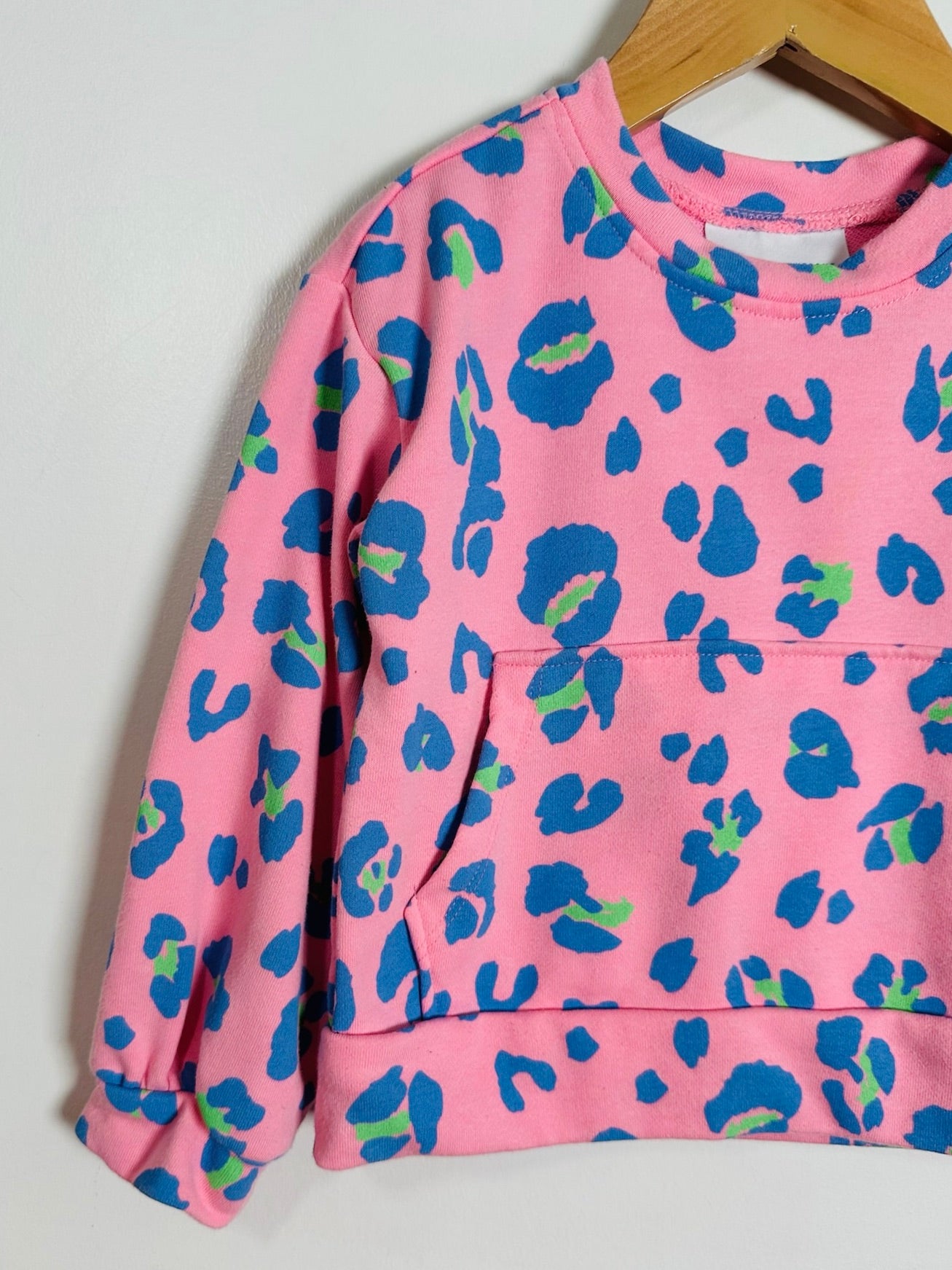 A pink long-sleeve sweatshirt with a blue and green abstract animal print, hanging on a wooden hanger against a plain light gray wall.