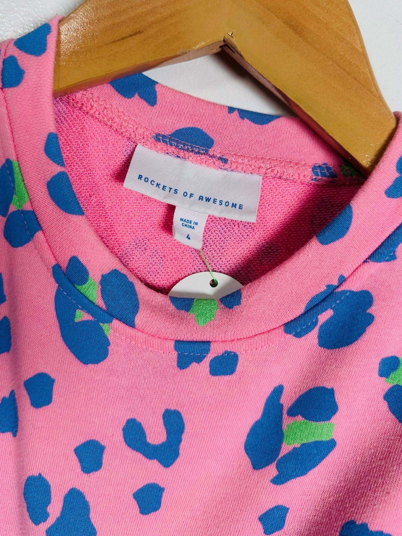 A close-up of a pink crewneck sweatshirt with a blue and green abstract pattern, hanging on a wooden hanger. The brand label reads 'ROCKETS OF AWESOME'.
