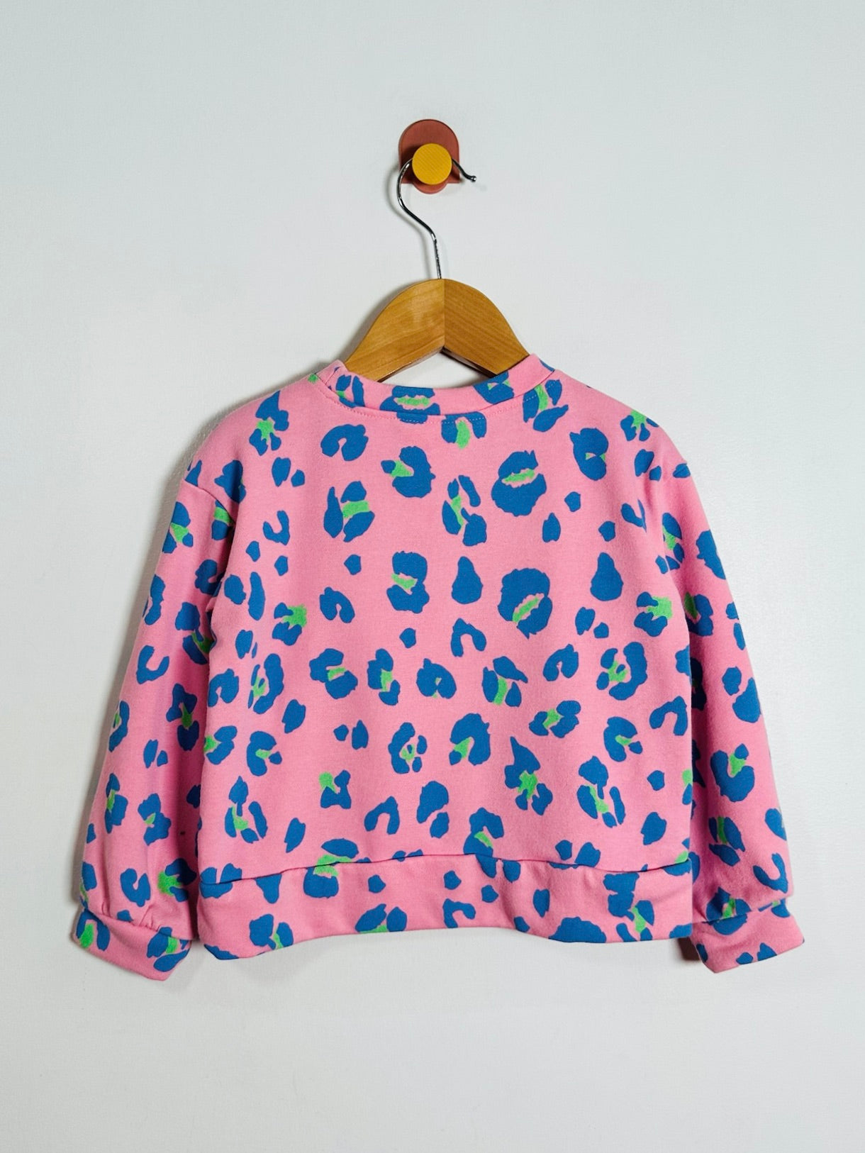 A pink long-sleeve sweatshirt with a blue and green leopard print pattern, hanging on a wooden hanger against a plain light gray wall.