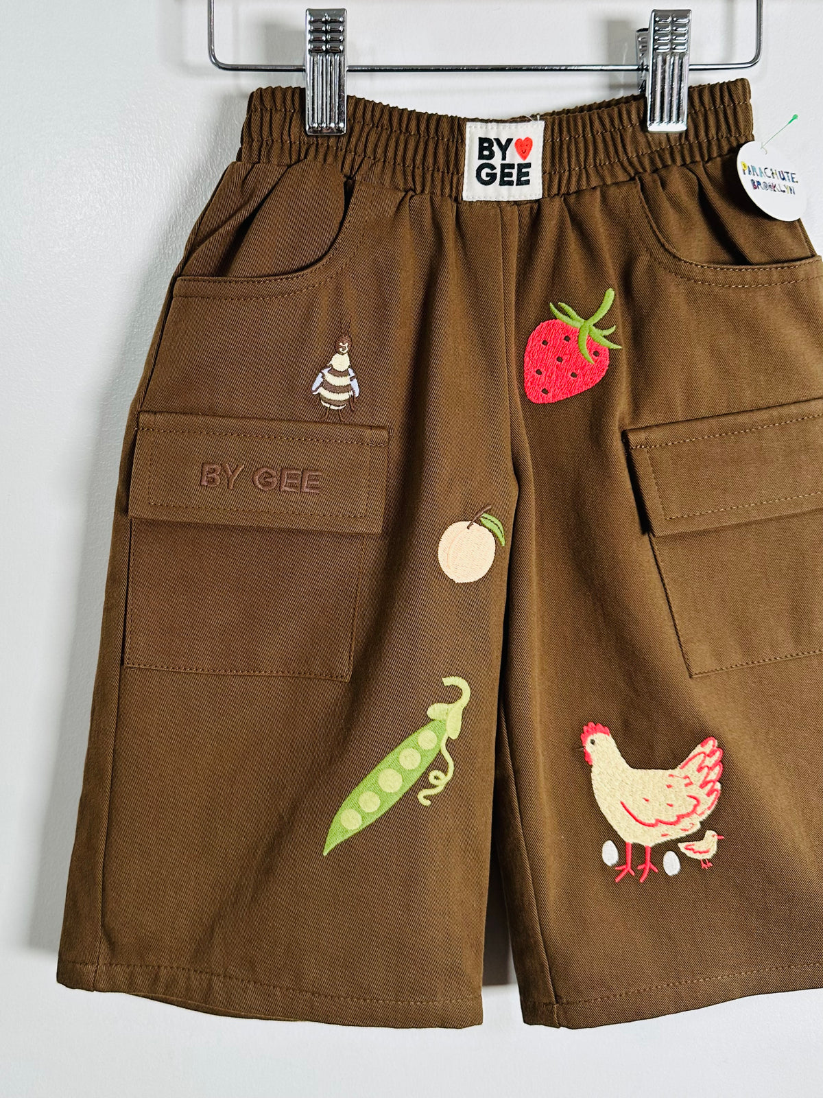 A pair of brown cargo shorts for kids by BY GEE, featuring embroidered vegetable and animal motifs including a strawberry, apple, pea pod, and chicken, hanging on a metal hanger against a plain white wall.