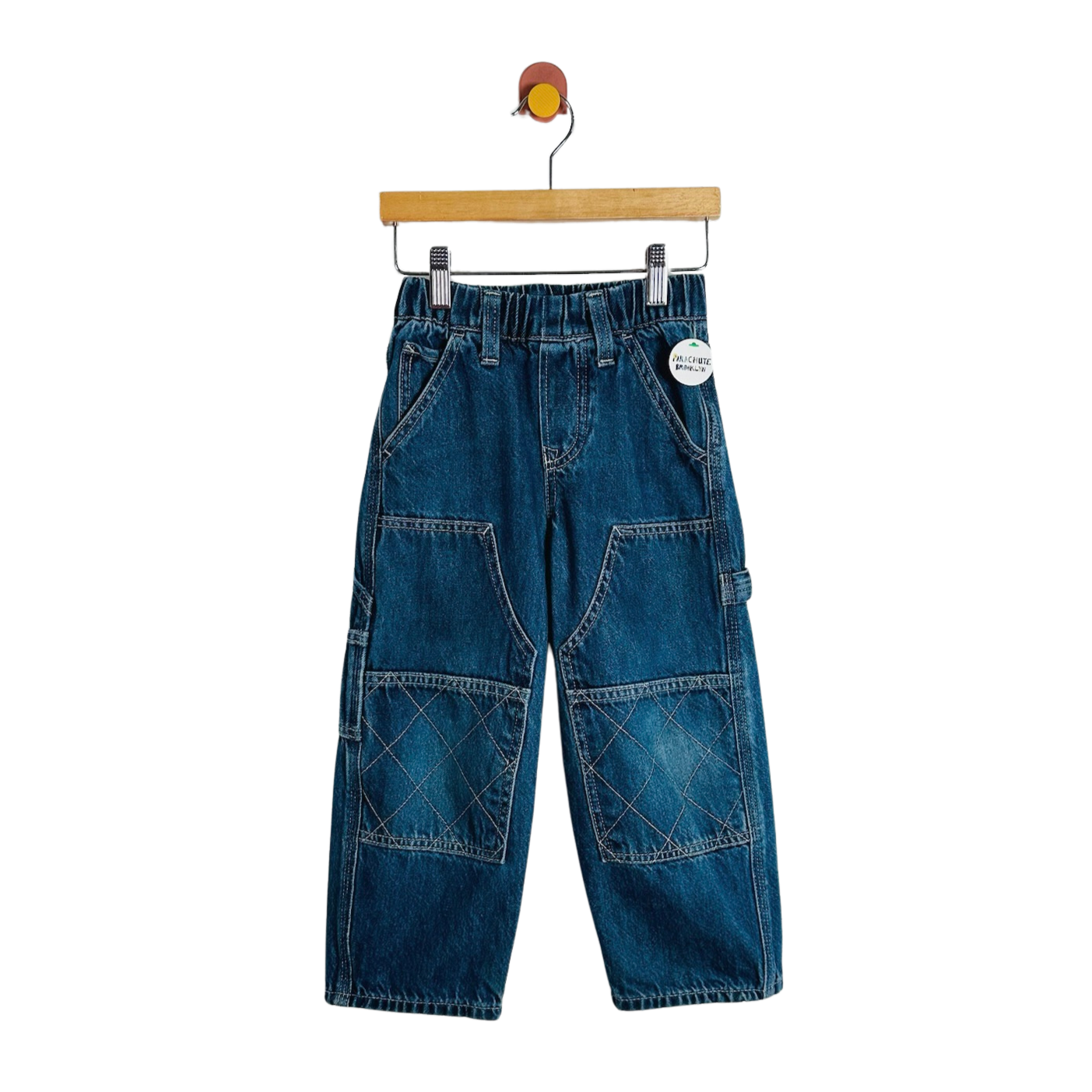 A pair of blue denim cargo pants for kids, hanging from a wooden hanger against a plain white background. The pants feature an elastic waistband, front patch pockets, and large diamond-stitched cargo pockets on the lower legs.
