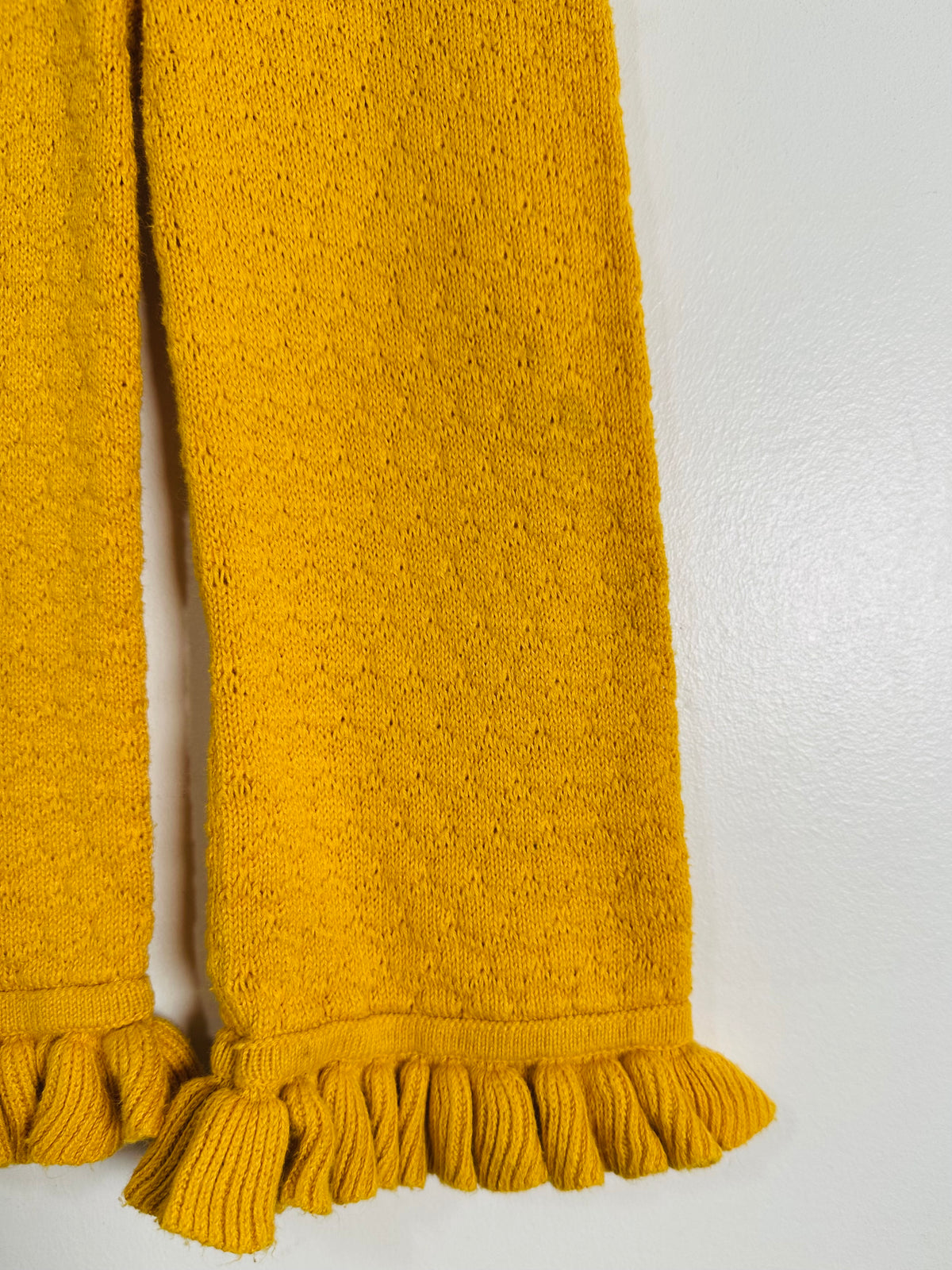 A close-up of a mustard yellow knit blanket or throw with fringed edges, laid against a plain light gray background.