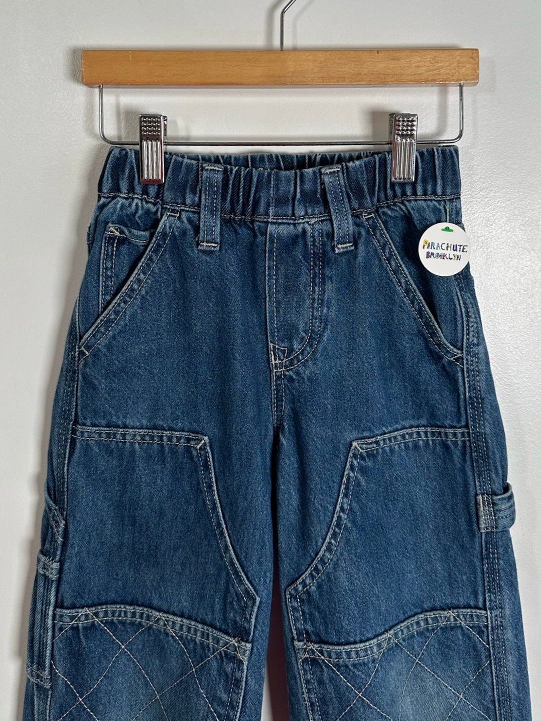 A pair of blue denim cargo pants hanging from a wooden hanger against a white wall. The pants feature an elastic waistband, front slash pockets, and large patch pockets on the thighs.
