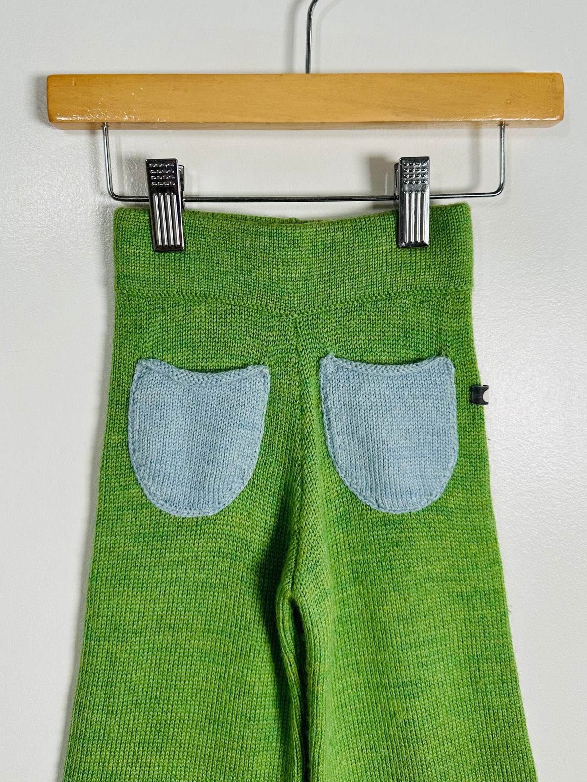 Green knit pants with two light blue patch pockets, hanging from a wooden hanger with metal clips against a plain light wall.