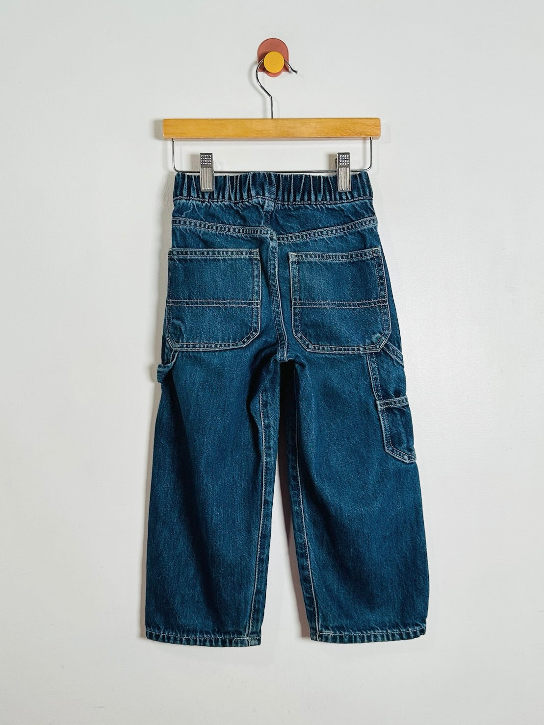 A pair of blue denim cargo pants shown from the back, hanging from a wooden hanger against a plain light gray wall.