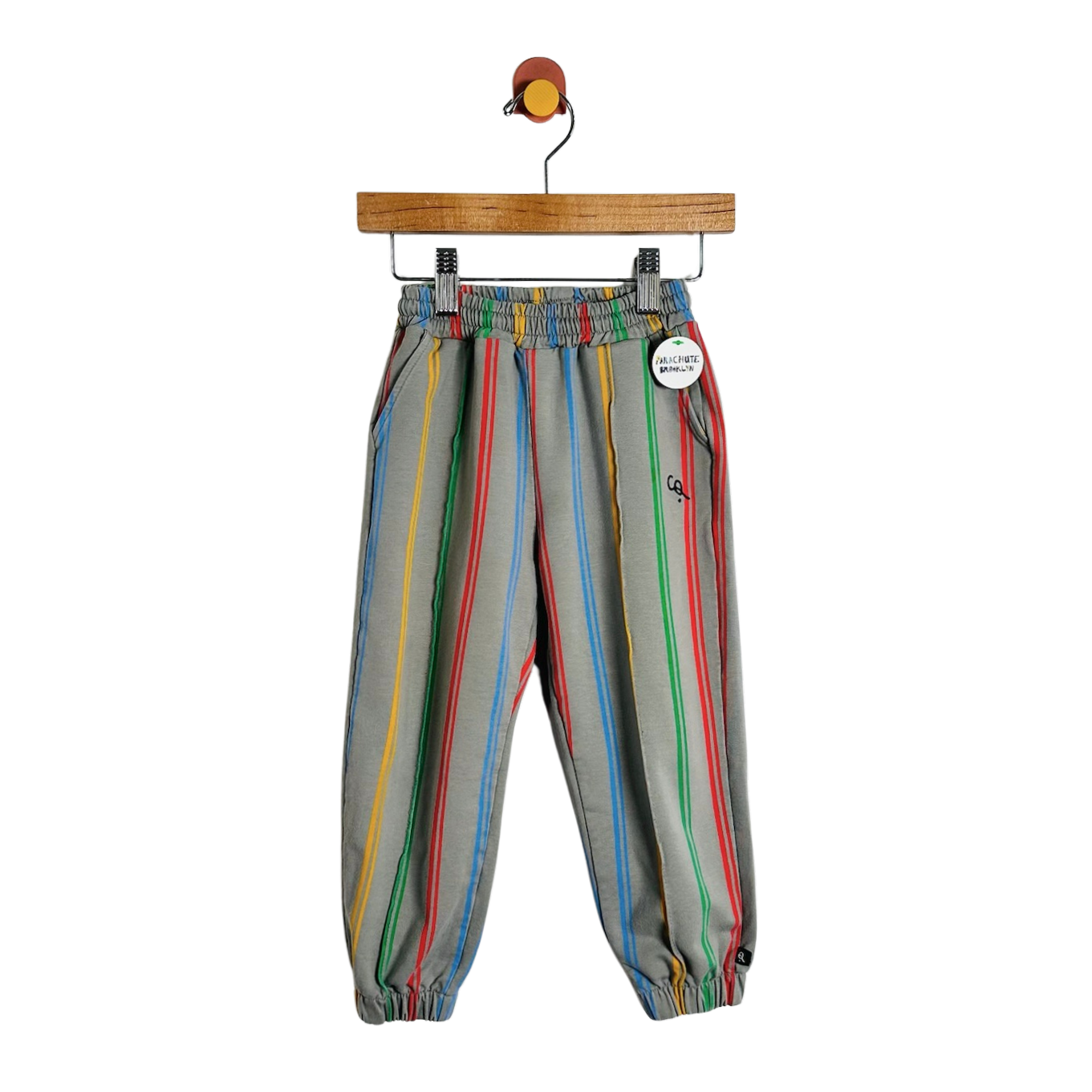 A pair of children's jogger pants with vertical rainbow stripes on a gray base, hanging from a wooden hanger against a plain white background.