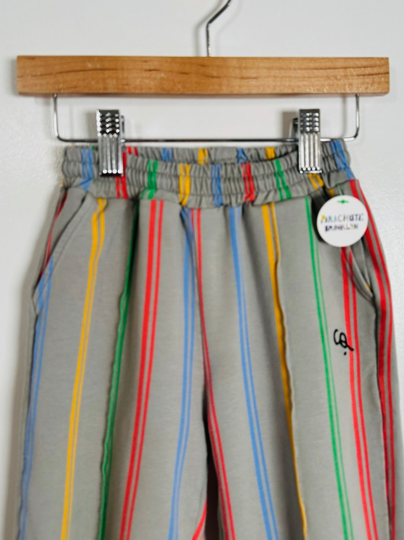 Gray joggers with vertical red, yellow, green, and blue stripes, hanging from a wooden hanger with metal clips against a white wall.