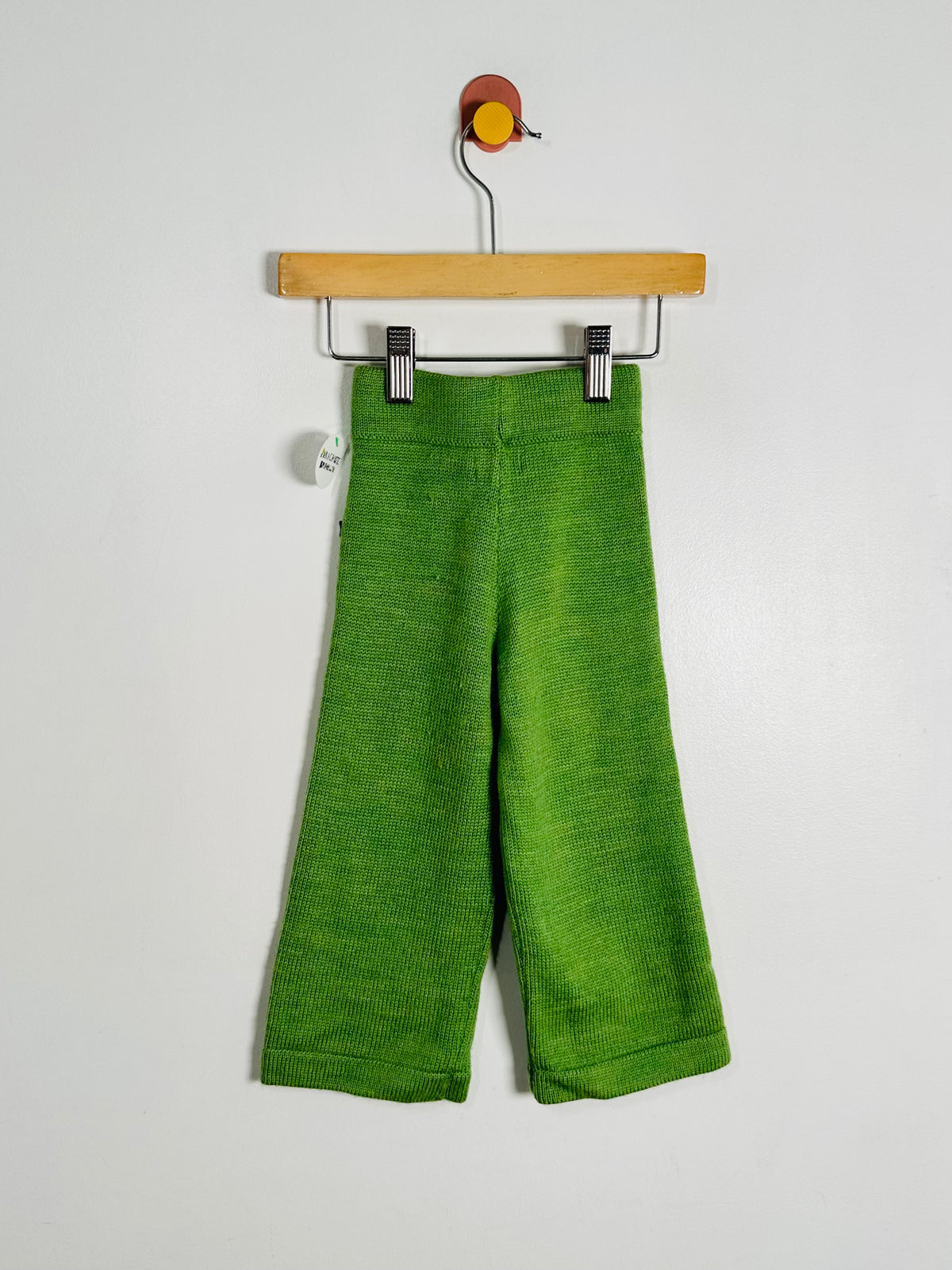 Bright green knit cropped pants hanging from a wooden hanger against a plain light gray wall.