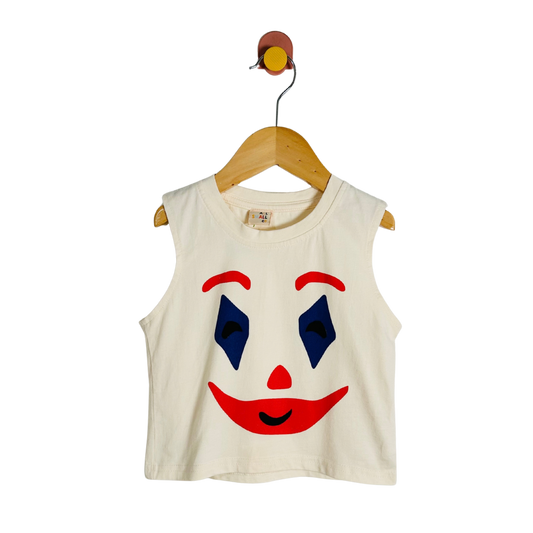 An off-white sleeveless crop top for kids, featuring a bold clown face graphic with red lips, a red nose, blue eyes, and red eyebrows. The shirt is displayed on a wooden hanger with a wall hook against a solid white background.