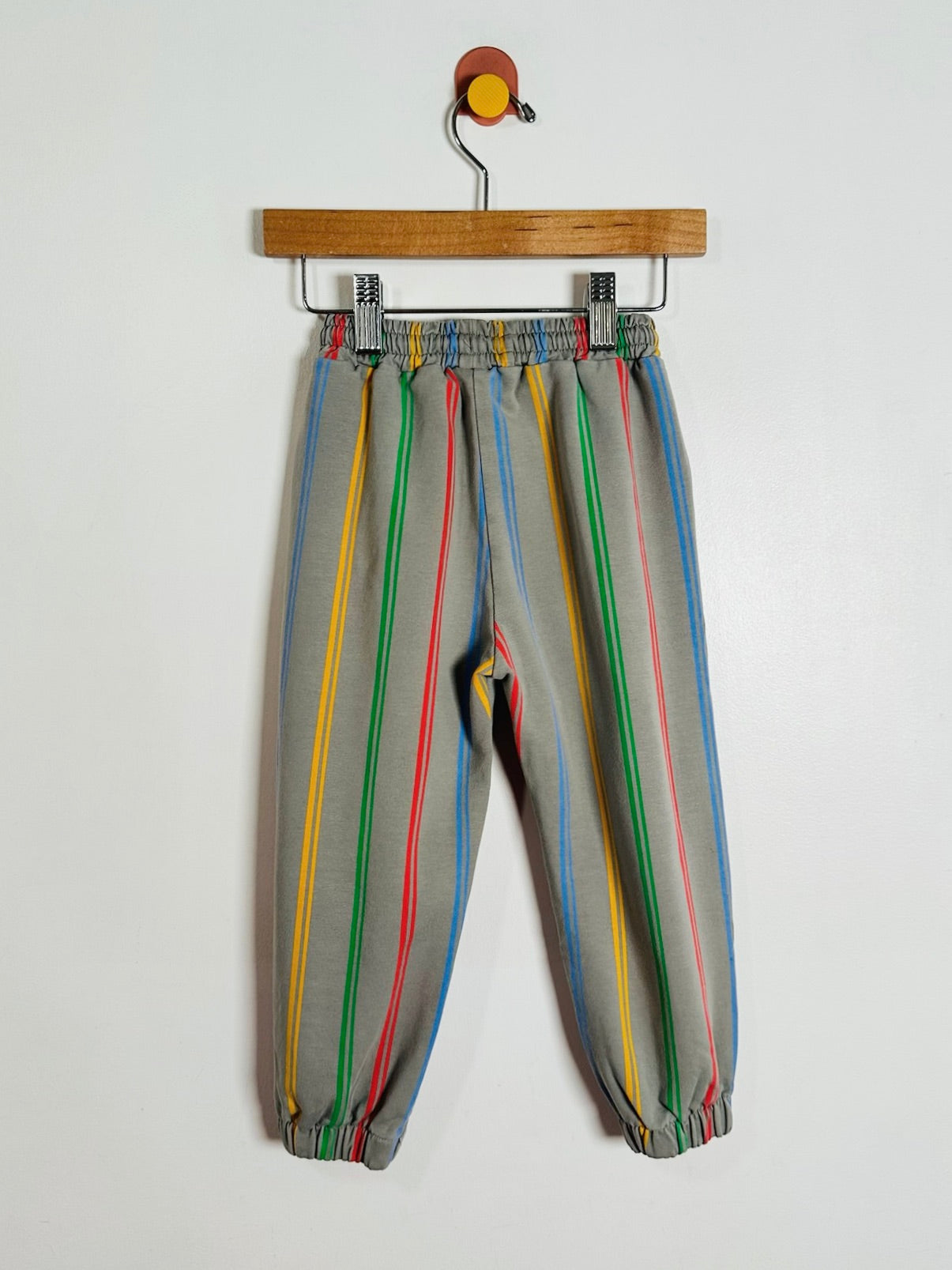A pair of gray jogger pants with vertical multicolor stripes in yellow, green, red, and blue, hanging from a wooden hanger against a plain white wall.
