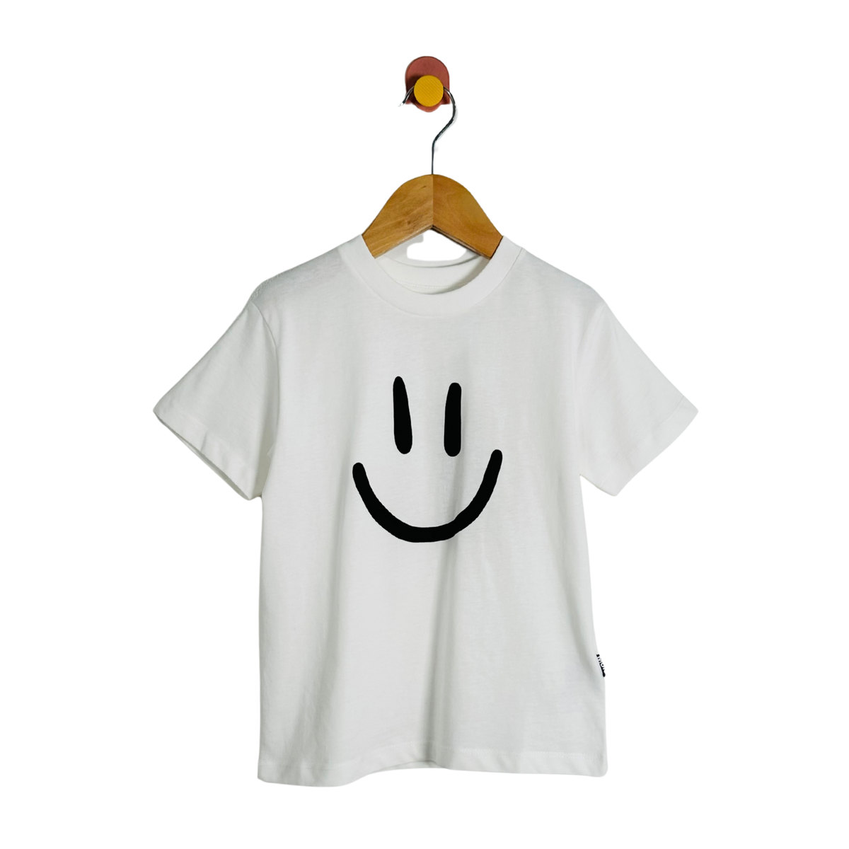 A white short-sleeve crewneck t-shirt featuring a large black smiley face graphic on the front, hanging on a wooden hanger from a wall hook against a solid white background.