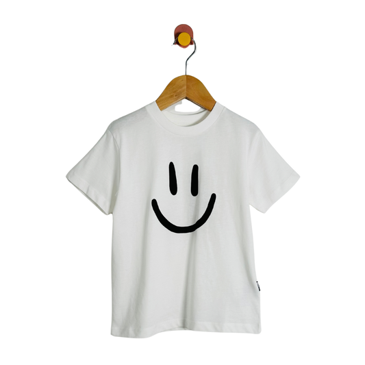A white short-sleeve crewneck t-shirt featuring a large black smiley face graphic on the front, hanging on a wooden hanger from a wall hook against a solid white background.